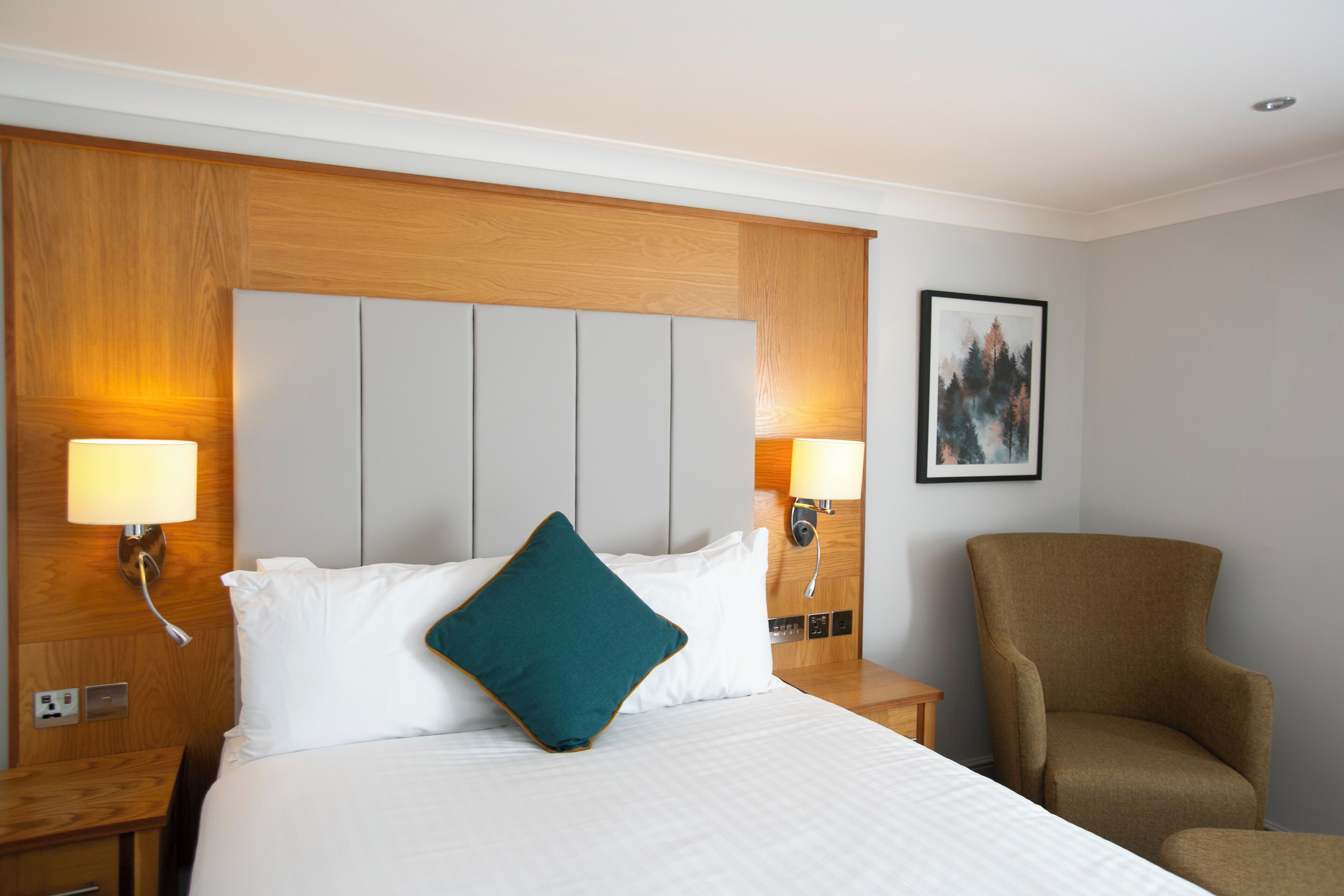 Ein Hotelzimmer mit einem großen Bett, zwei Nachttischlampen, einem hölzernen Kopfteil, einem Sessel und einem gerahmten Bild an der Wand.