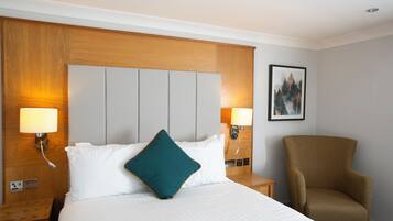 Ein Hotelzimmer mit einem großen Bett, zwei Nachttischlampen, einem hölzernen Kopfteil, einem Sessel und einem gerahmten Bild an der Wand.