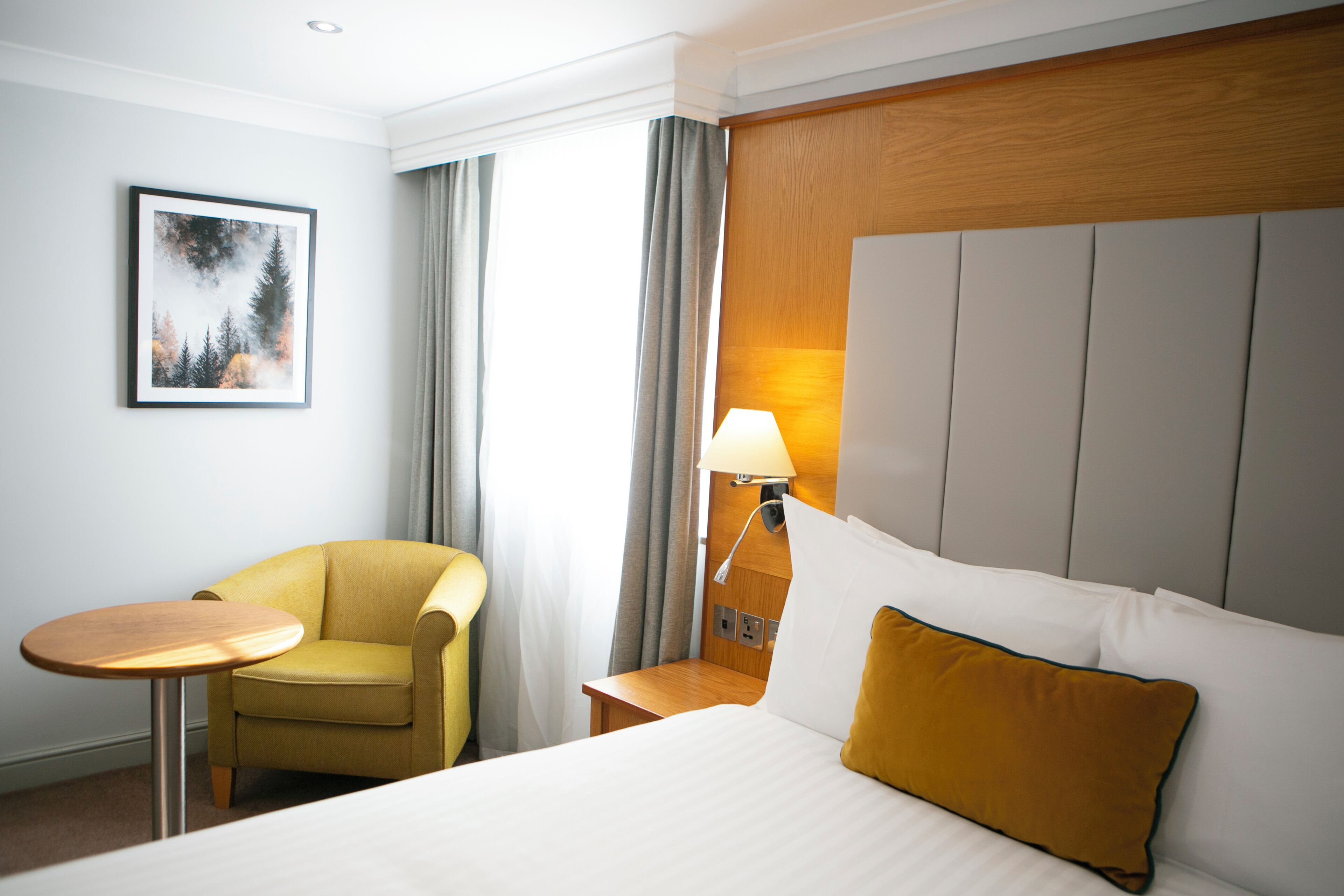 Una habitación de hotel con una cama, una silla, una mesita, una mesita de noche, una lámpara y un cuadro en la pared.