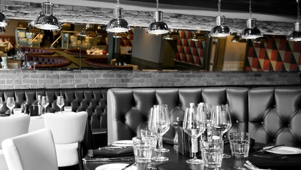 A restaurant with a black leather booth seating area and white chairs. Tables are set with wine glasses, water glasses, and cutlery. - Crowne Plaza East Midlands Airport by IHG (Kegworth)