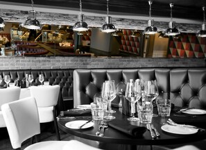 A restaurant with a black leather booth seating area and white chairs. Tables are set with wine glasses, water glasses, and cutlery. - Crowne Plaza East Midlands Airport by IHG (Kegworth)