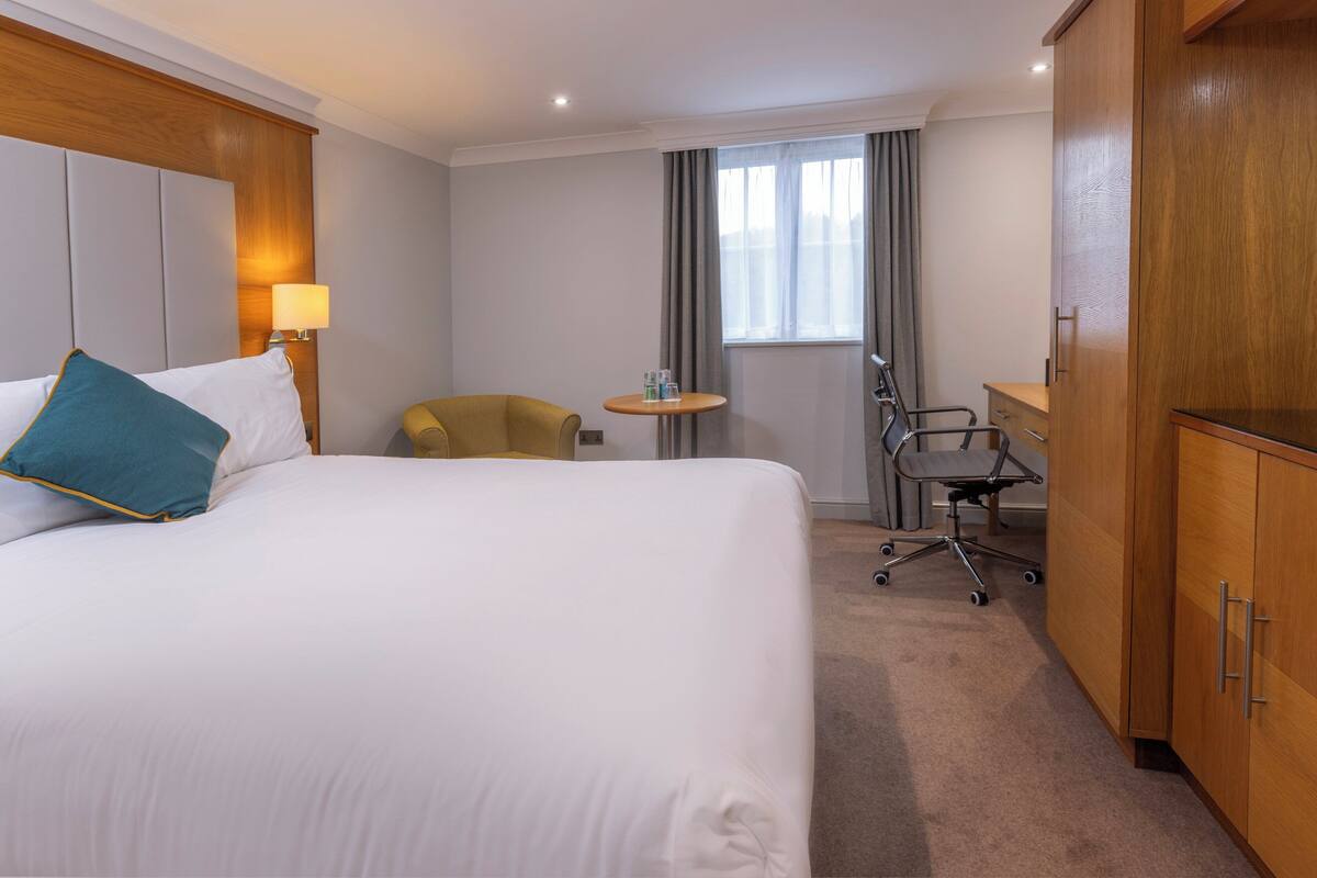 A hotel room with a large bed, a desk, a chair, a wardrobe, and a small table with a lamp.