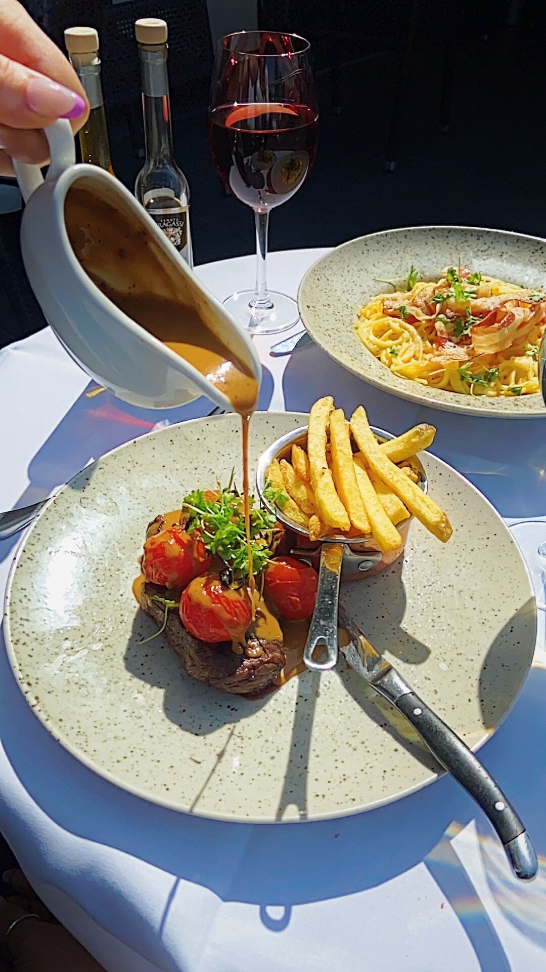 A meal with steak, fries, and sauce on a plate, with a wine glass and bottles in the background.