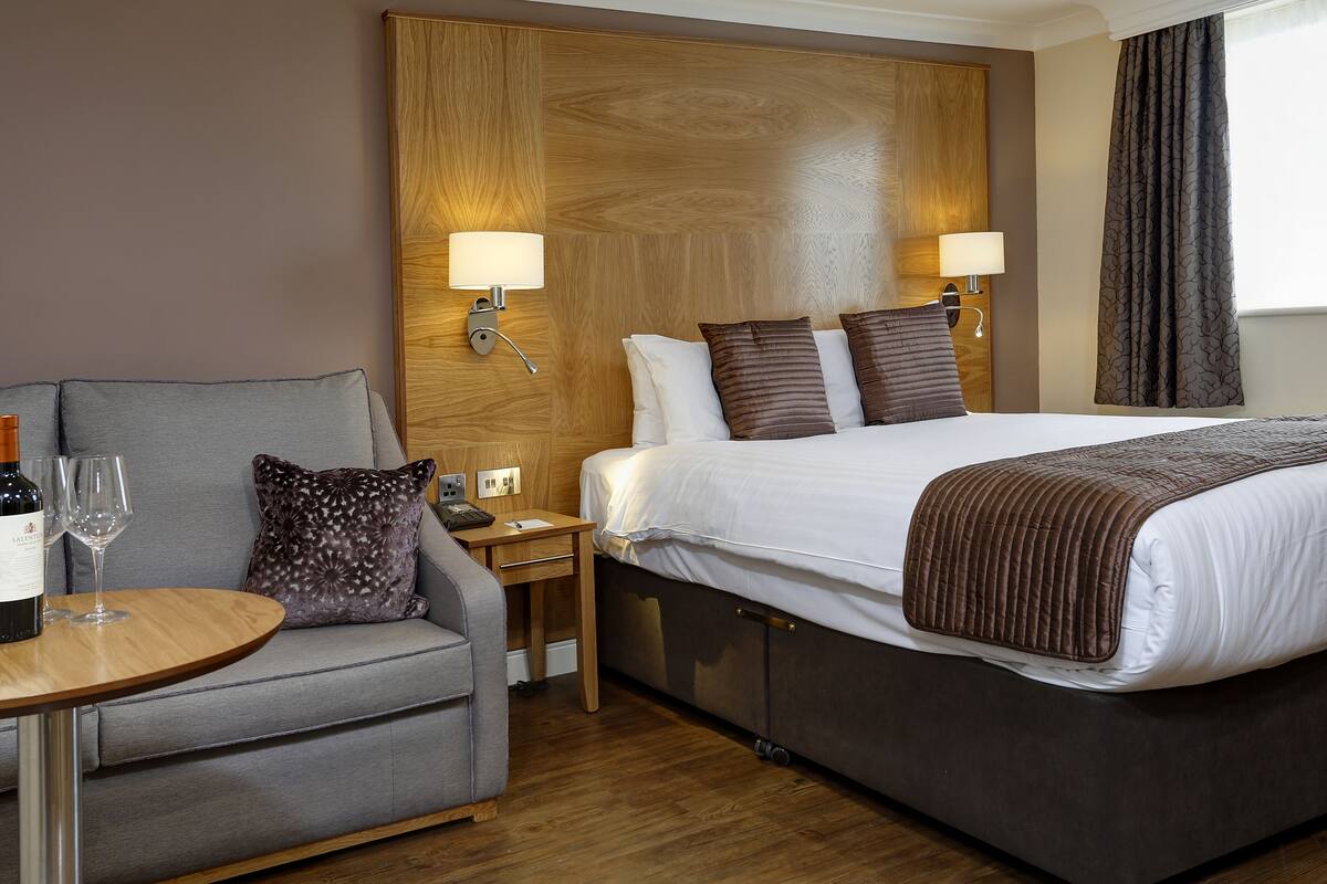 A hotel room with a bed, a sofa, a small table with wine glasses, and a nightstand.