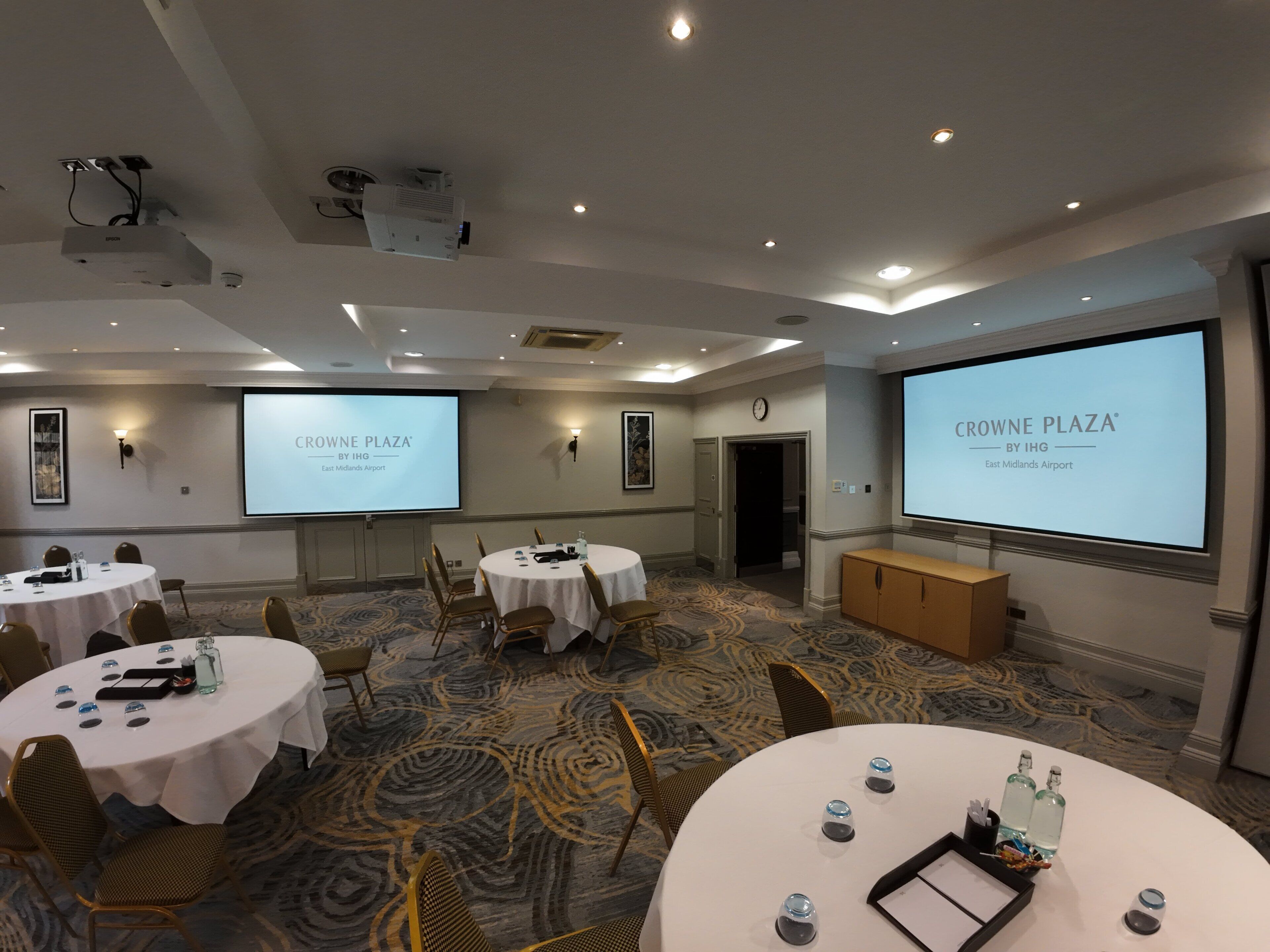 A conference room with multiple tables, chairs, and presentation screens displaying "CROWNE PLAZA BY IHG East Midlands Airport".