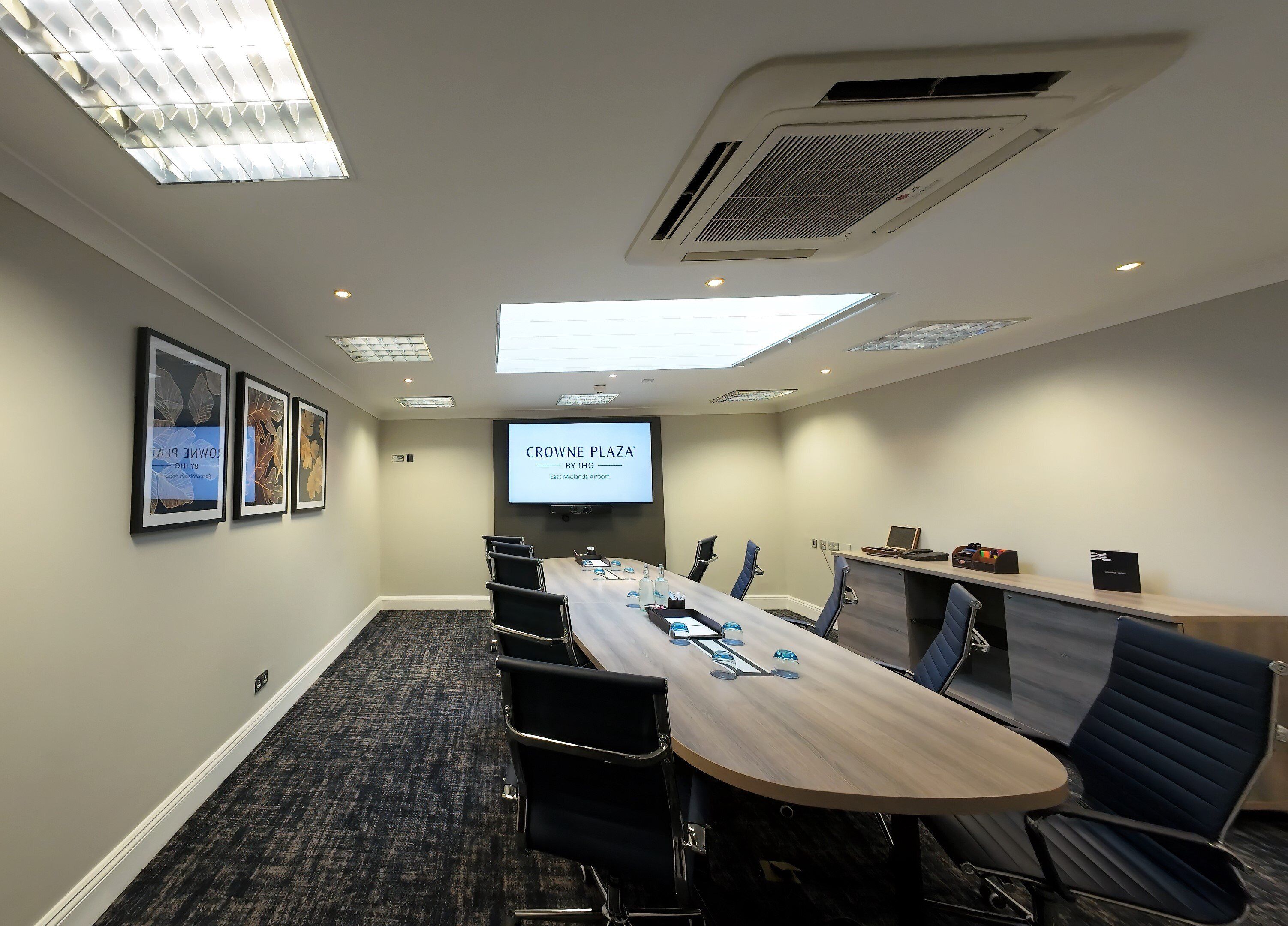 A conference room with a long table, chairs, a projector screen displaying "CROWNE PLAZA BY IHG East Midlands Airport", and framed artwork on the wall.