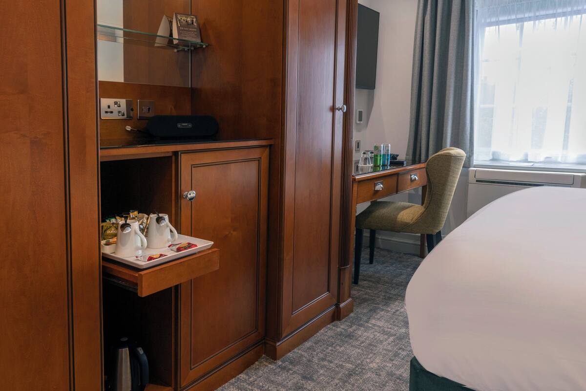 A hotel room with a wooden wardrobe, a desk with a chair, a bed, and a window with curtains.