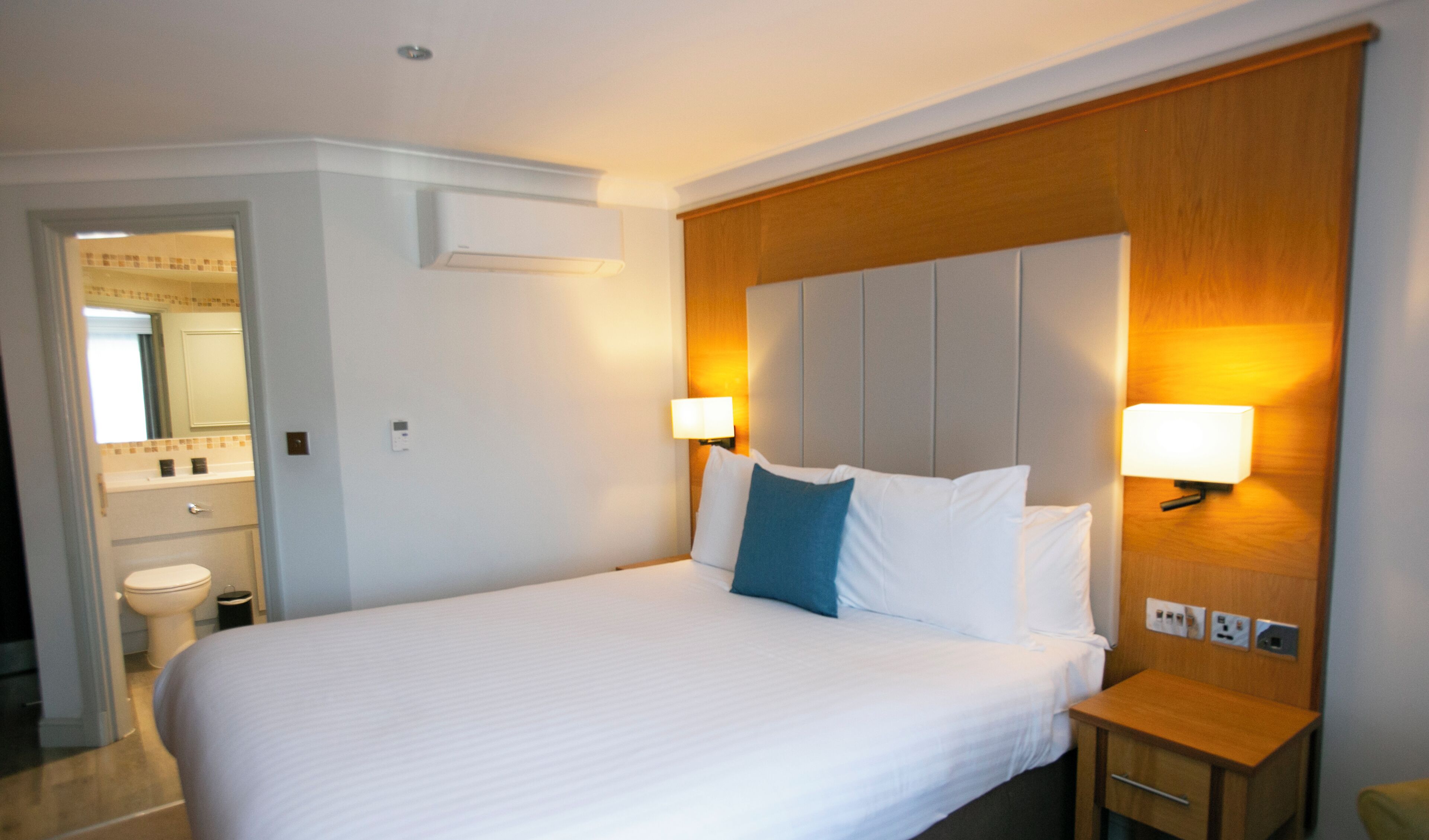 A hotel room with a large bed, bedside lamps, a wooden headboard, a nightstand, and a bathroom visible in the background.