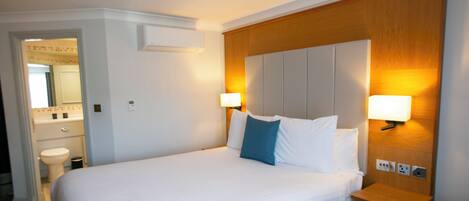 A hotel room with a large bed, bedside lamps, a wooden headboard, a nightstand, and a bathroom visible in the background.
