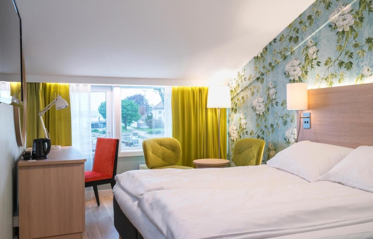 Een hotelkamer met een bed, twee stoelen, een bureau met een lamp, een klein tafeltje en een raam met gordijnen.