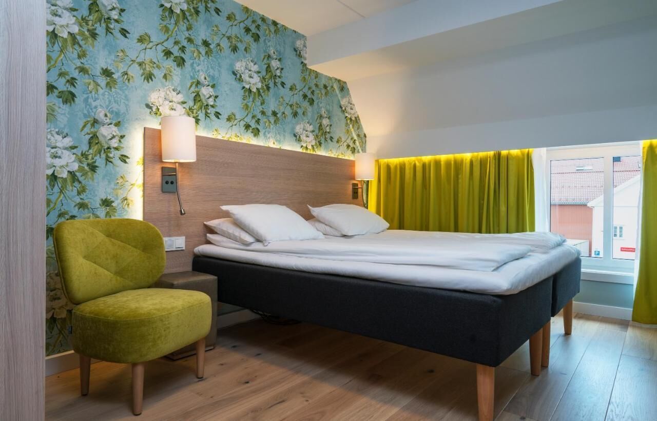 Ein Schlafzimmer mit einem Bett, einem Nachttisch, einer Lampe, einem Sessel und einem Fenster mit Vorhängen.