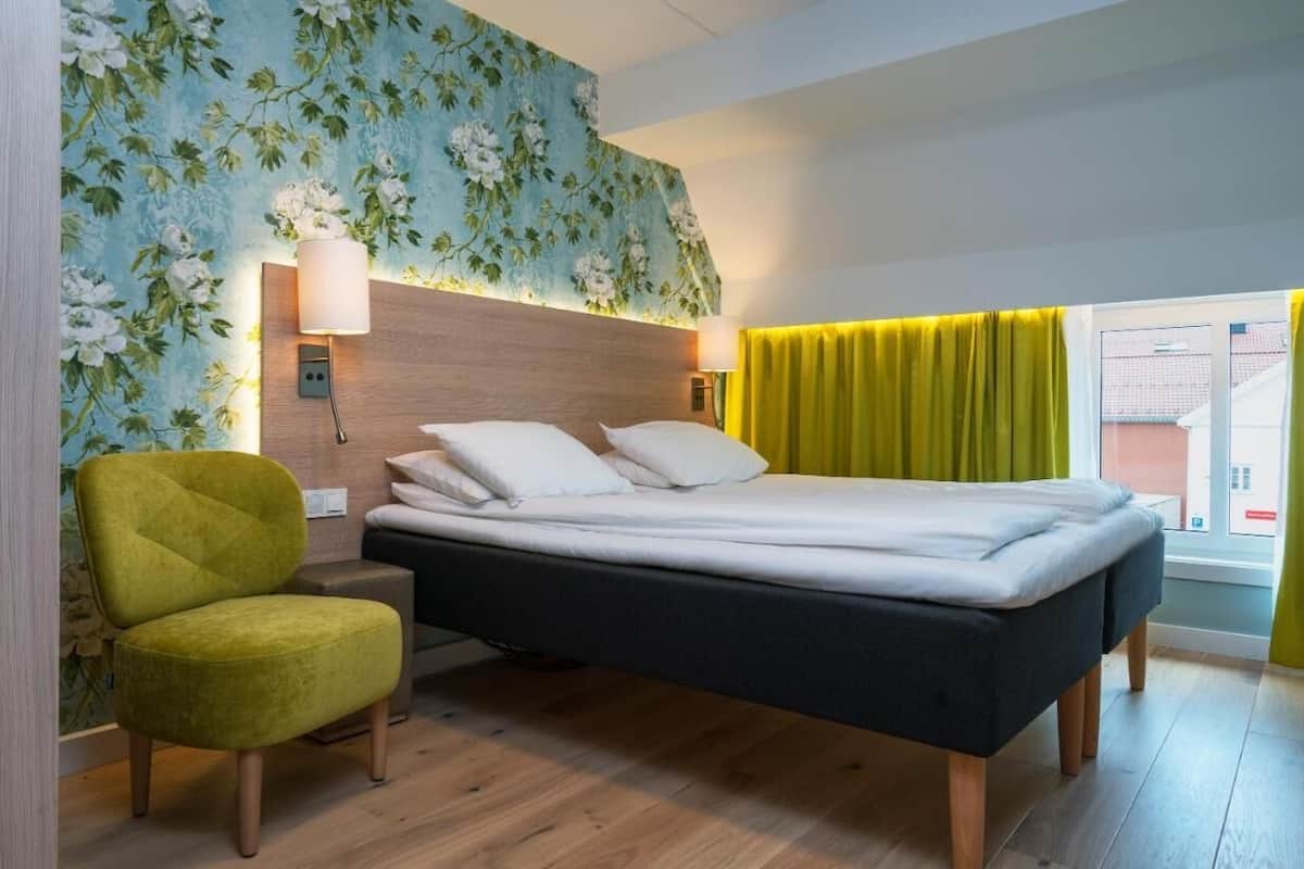 Een slaapkamer met een bed, nachtkastje, lamp, stoel en raam met gordijnen.