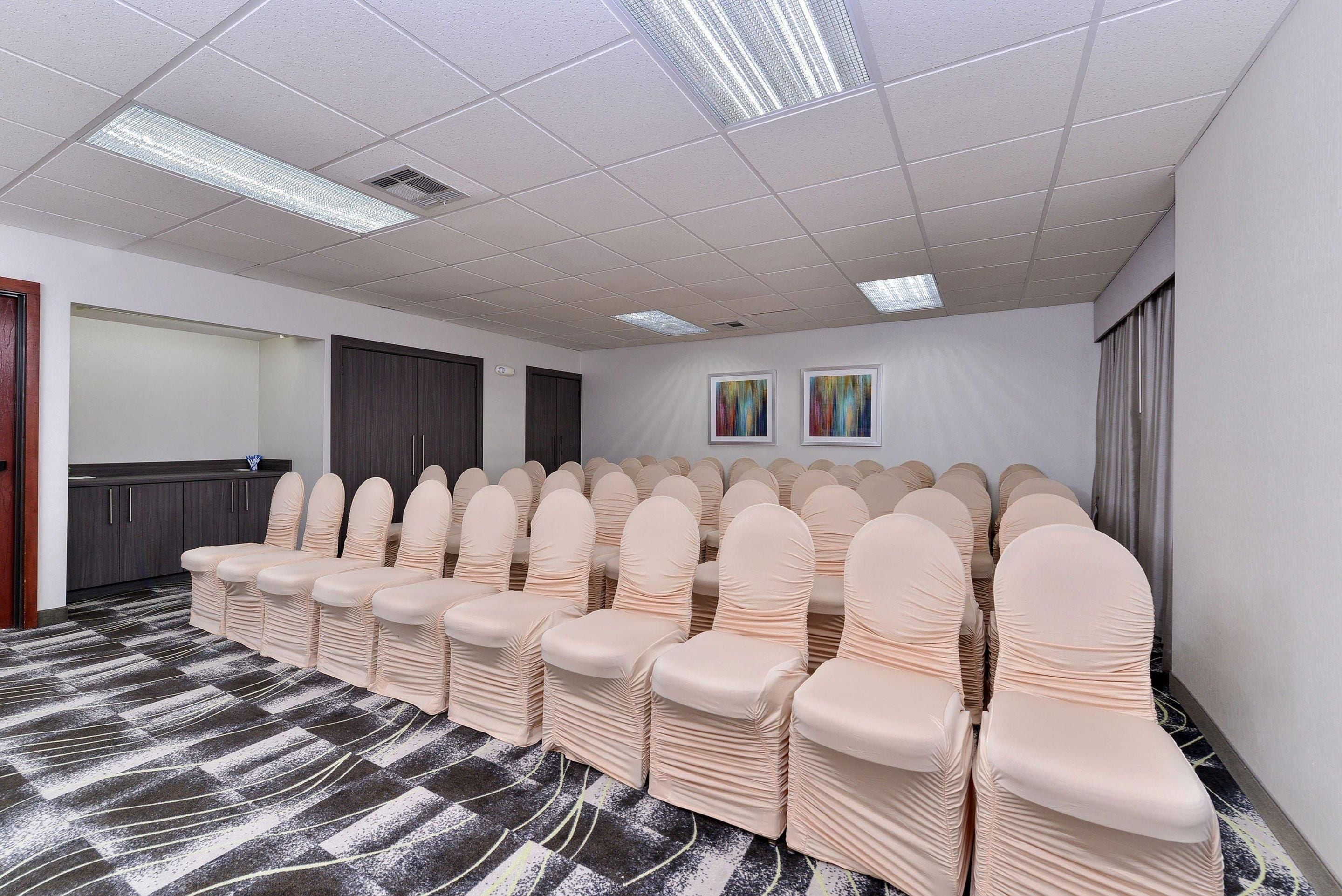 A conference room with rows of chairs covered in beige chair covers, a carpeted floor, a white ceiling with fluorescent lights, and two framed artworks on the wall.