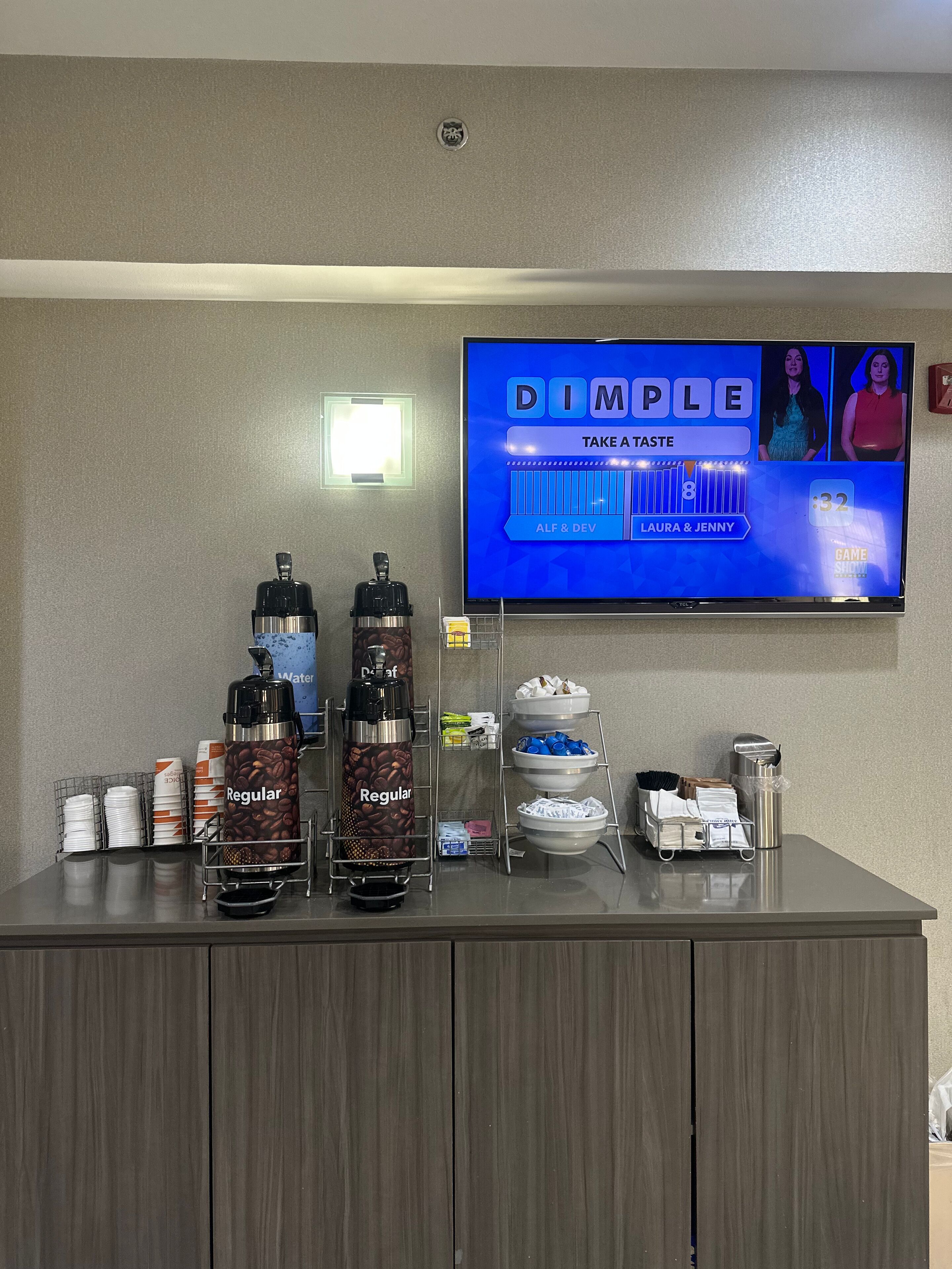 A reception area with a coffee station, a television displaying 'DIMPLE TAKE A TASTE', and a clock showing 32 minutes.