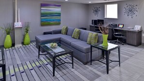 A modern living room with a gray sectional sofa, a glass coffee table, and a desk with a computer. There are decorative vases and a colorful abstract painting on the wall. - Comfort Inn & Suites Frisco - Plano (Frisco)