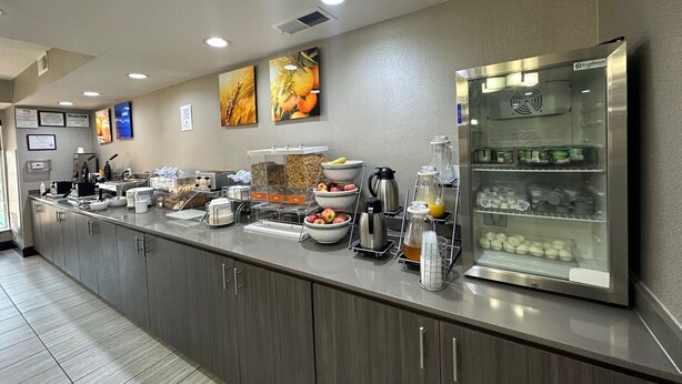 A modern kitchen area with a stainless steel counter, various food items, a coffee maker, and a refrigerator. - Comfort Inn & Suites Frisco - Plano (Frisco)