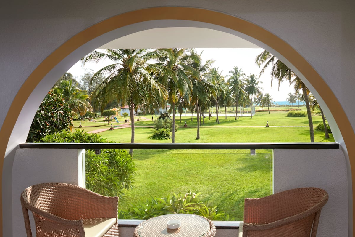 A balcony with wicker furniture and a glass table, overlooking a well-maintained lawn and palm trees.