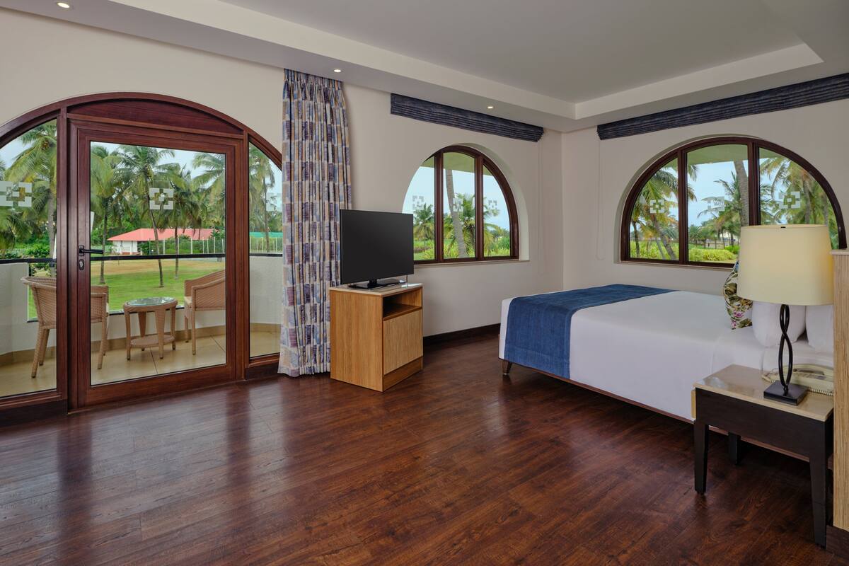 A hotel room with a wooden floor, a bed with a blue blanket, a TV on a wooden stand, a desk with a lamp, and large arched windows with a view of palm trees.