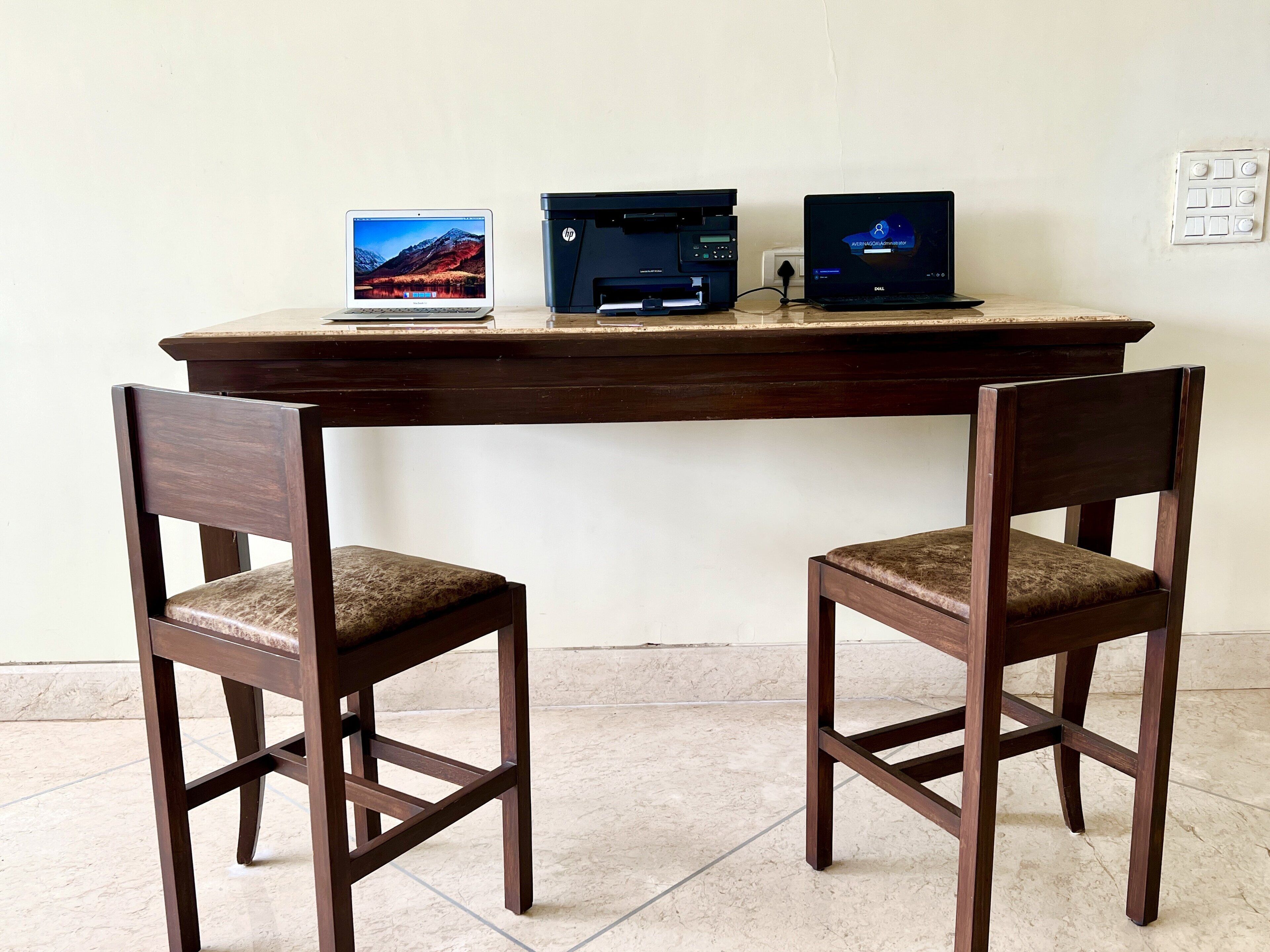 A wooden desk with two laptops and a printer, flanked by two wooden chairs with cushioned seats.