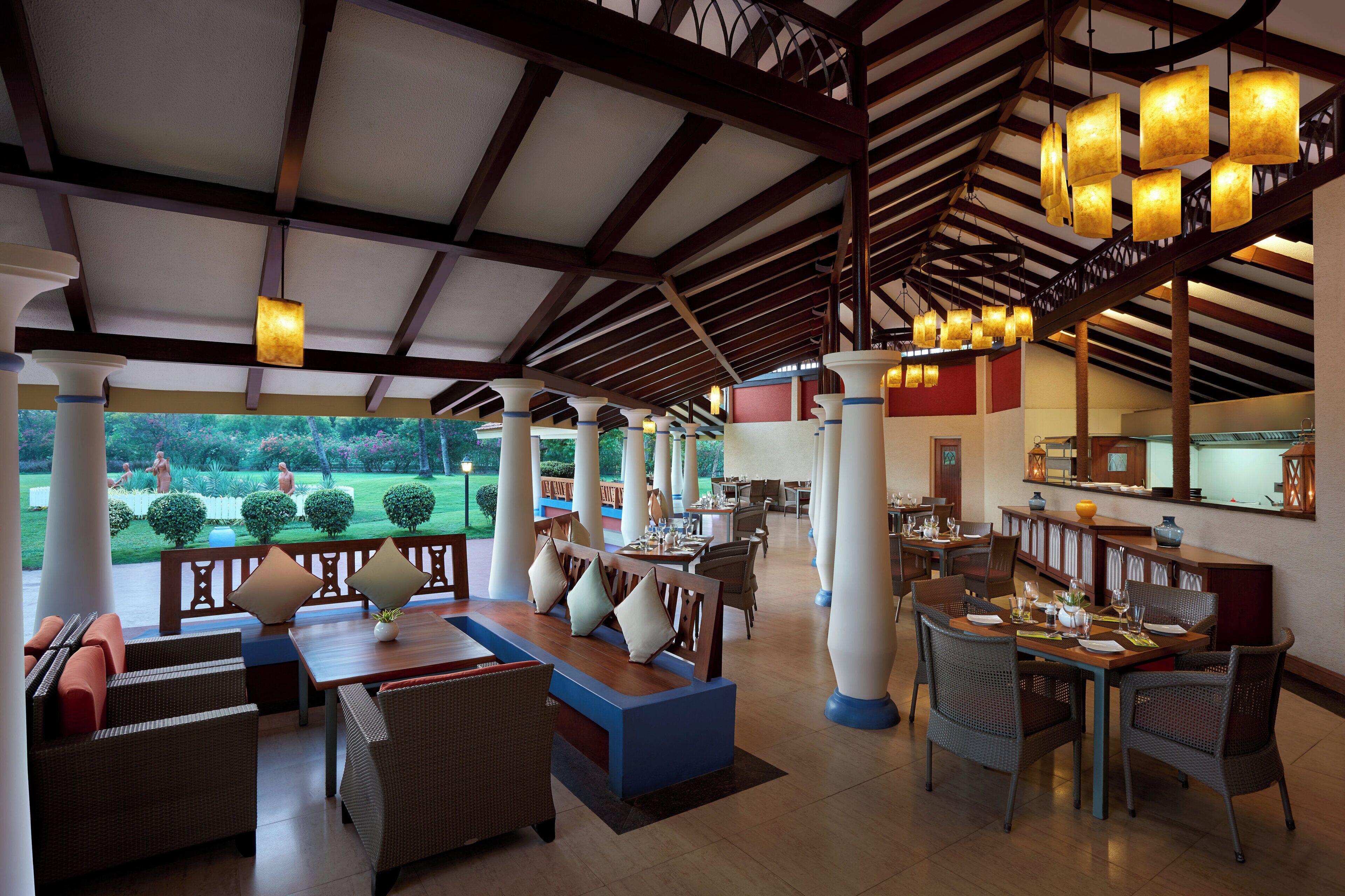 A spacious outdoor dining area with multiple tables and chairs, a reception desk, and a view of a landscaped garden.