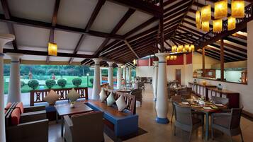 A spacious outdoor dining area with multiple tables and chairs, a reception desk, and a view of a landscaped garden.