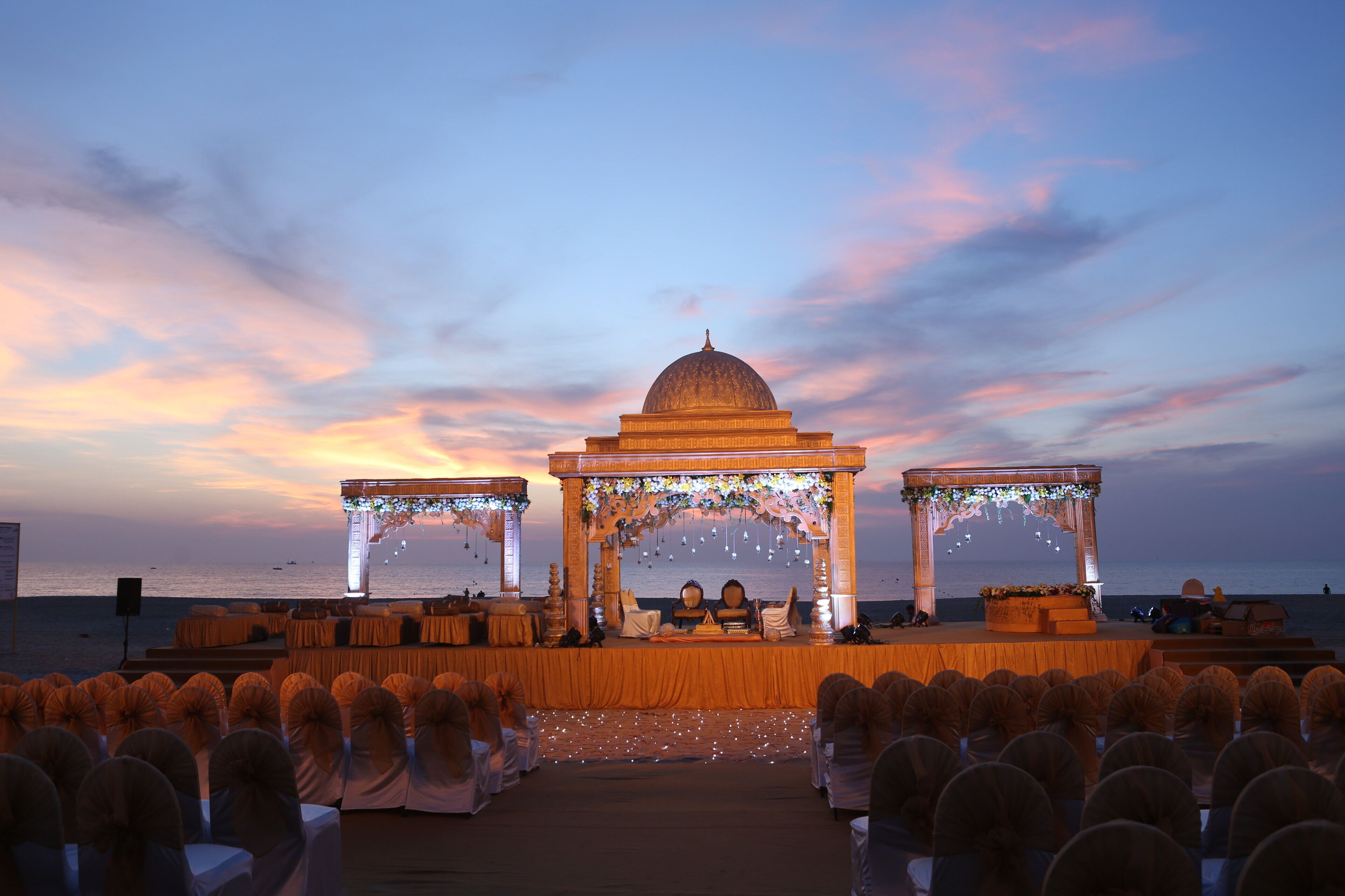 An outdoor wedding setup with a stage, seating, and a backdrop of a sunset over the sea.