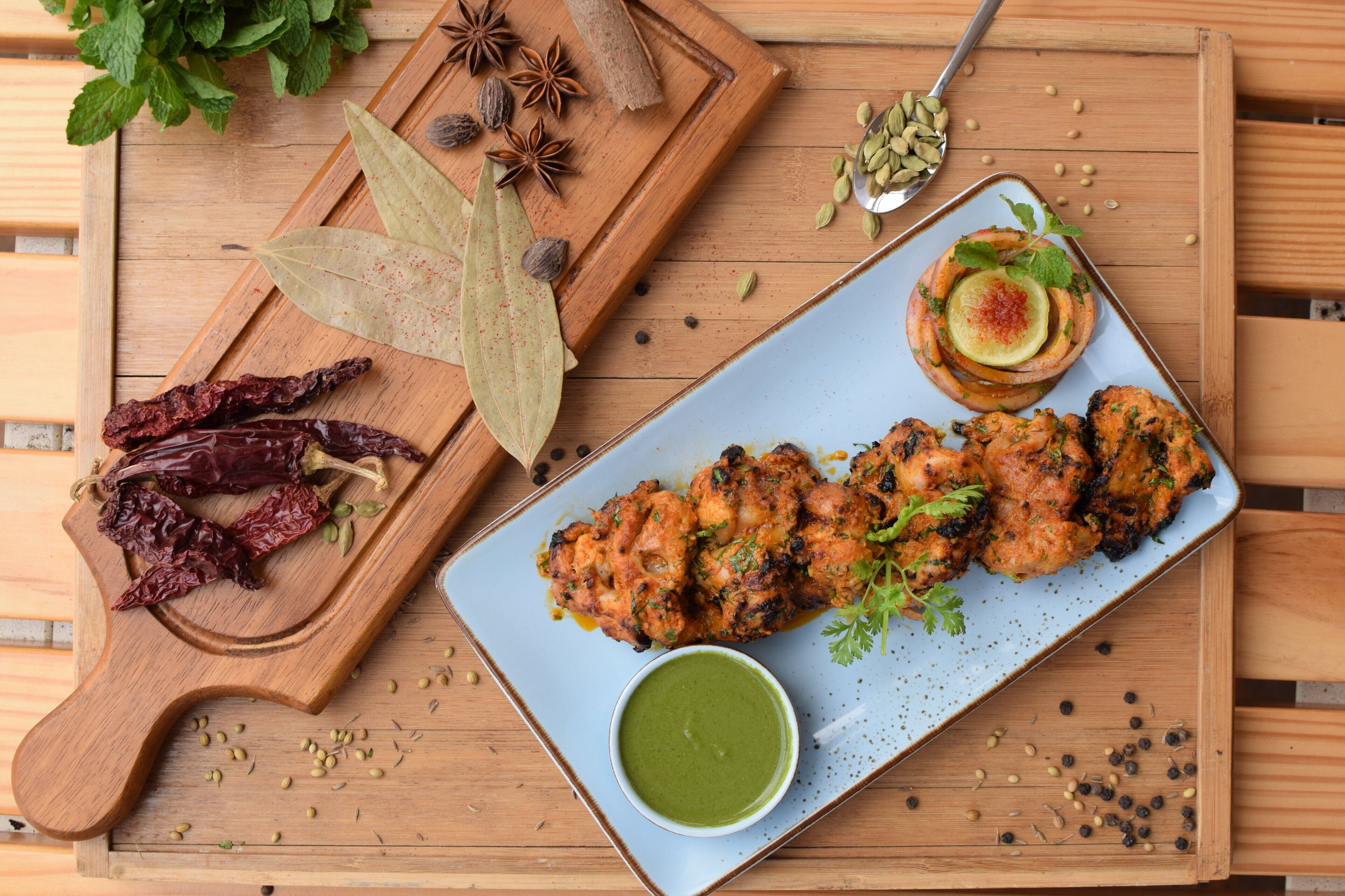 Une table en bois sur laquelle se trouvent une assiette de poulet grillé, un bol de sauce verte et une planche en bois avec des épices et des piments séchés.