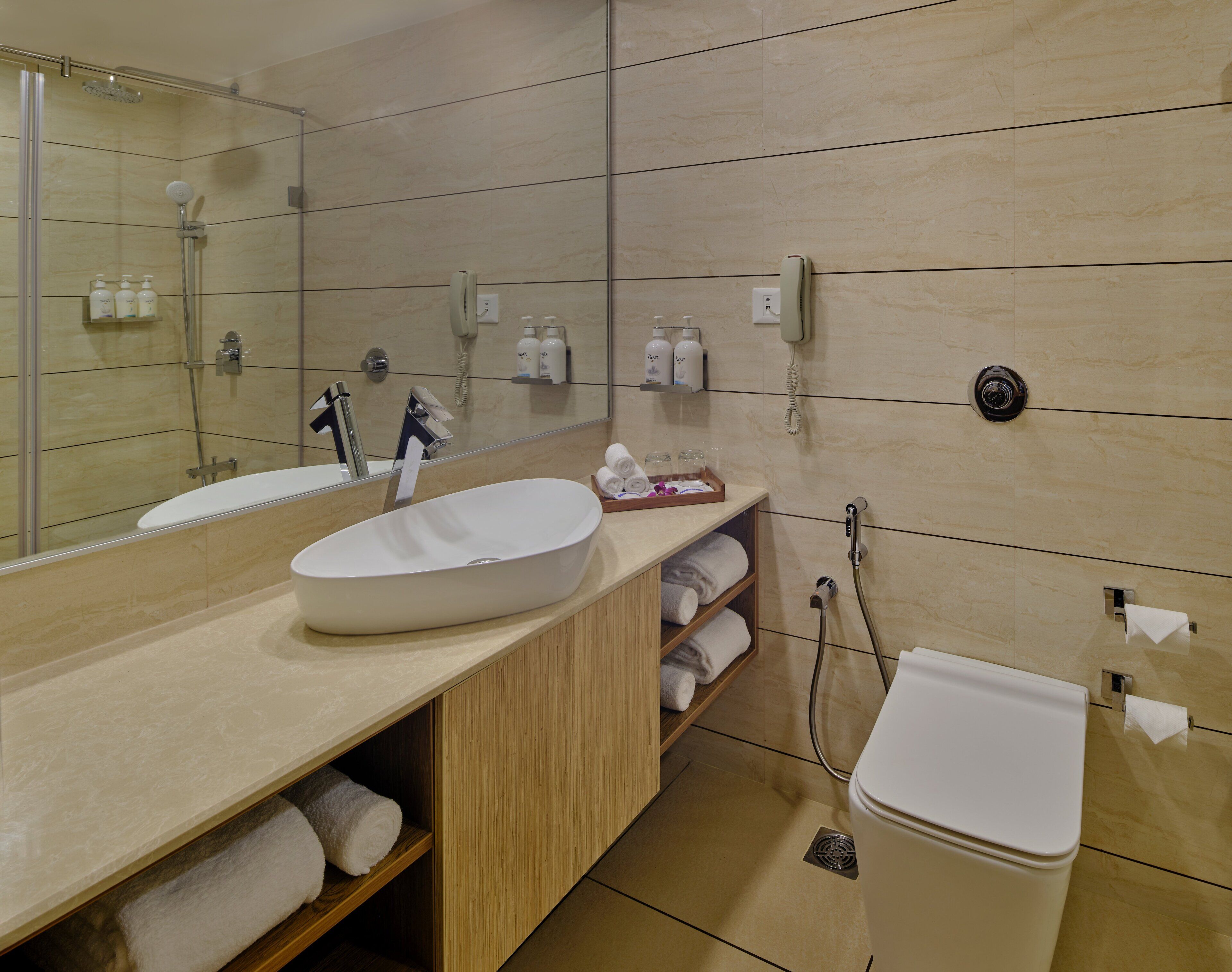 A modern bathroom with a large mirror, two sinks, a shower, and a toilet. There are towels and toiletries on the counter.