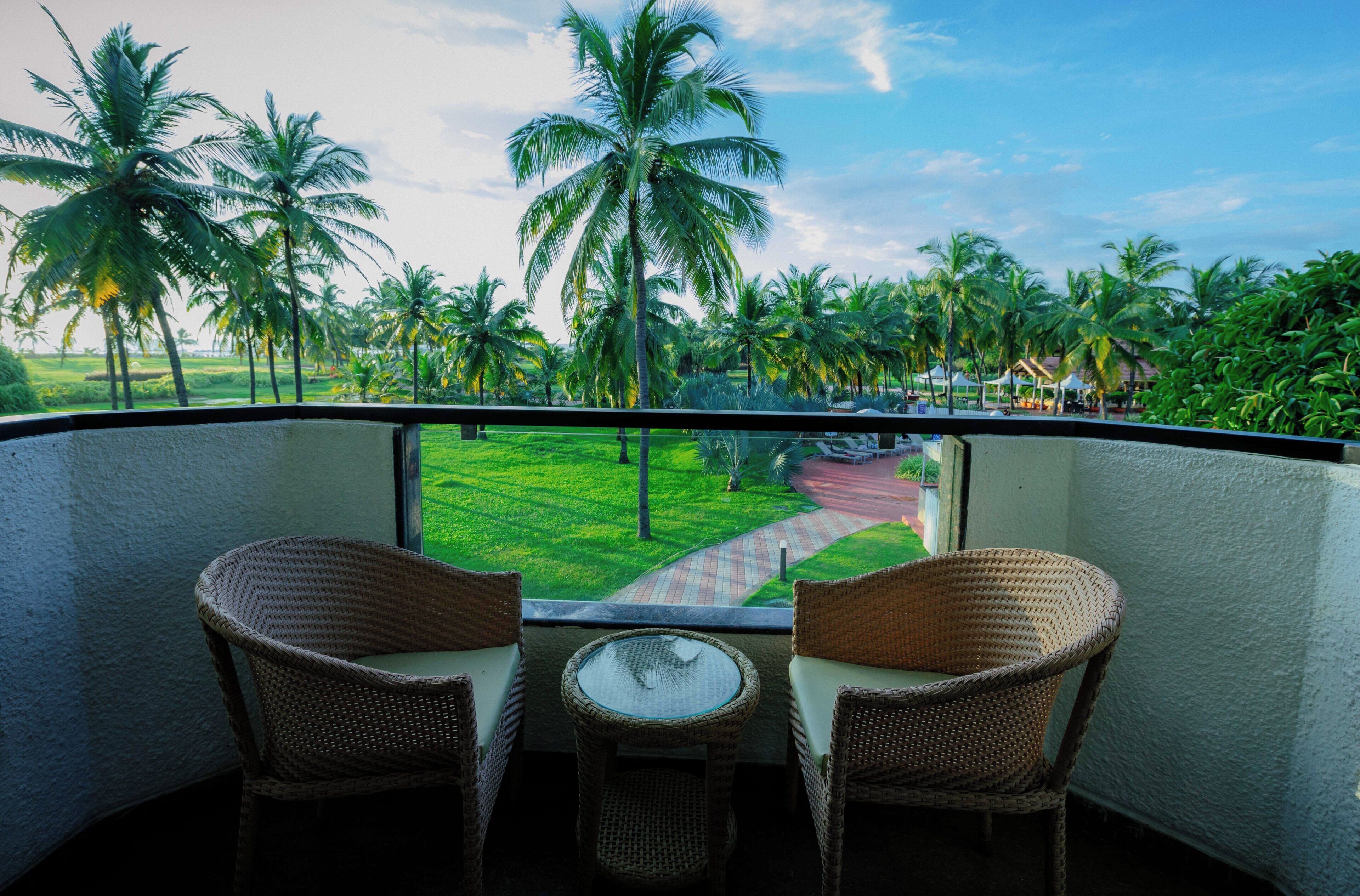 A balcony with wicker chairs and a small table, overlooking a lush landscape with palm trees and a pathway.