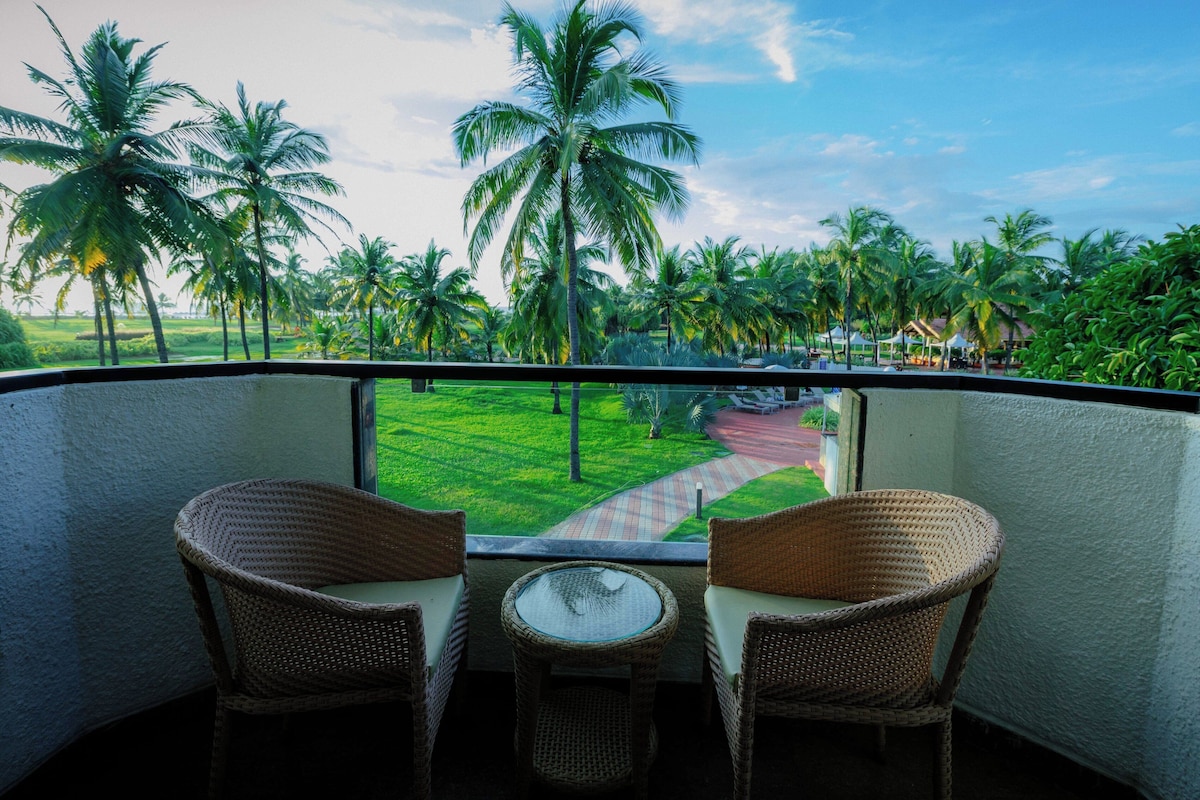 A balcony with wicker chairs and a small table, overlooking a lush garden with palm trees and a pathway.
