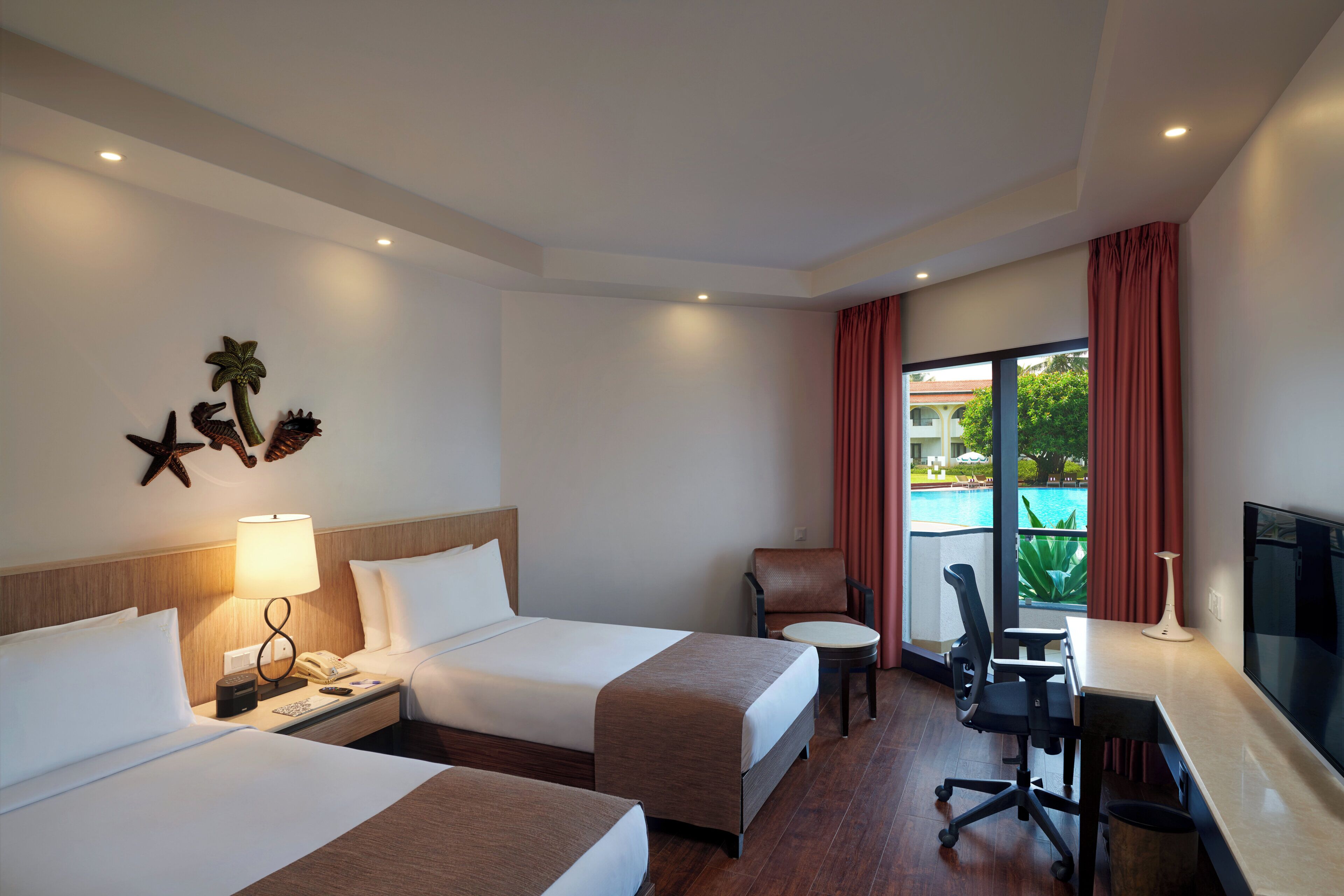 A hotel room with two beds, a desk, a chair, a TV, and a window with a view of a pool.