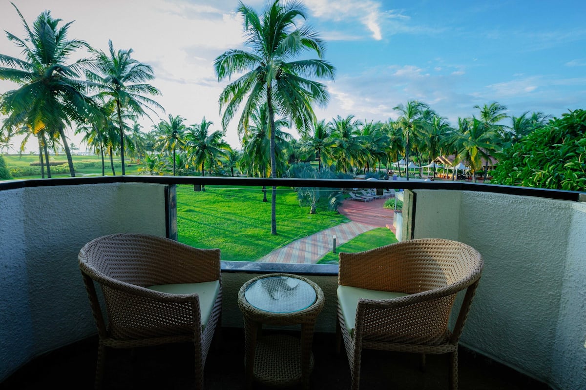 A balcony with wicker chairs and a small table, overlooking a lush garden with palm trees and a pathway.
