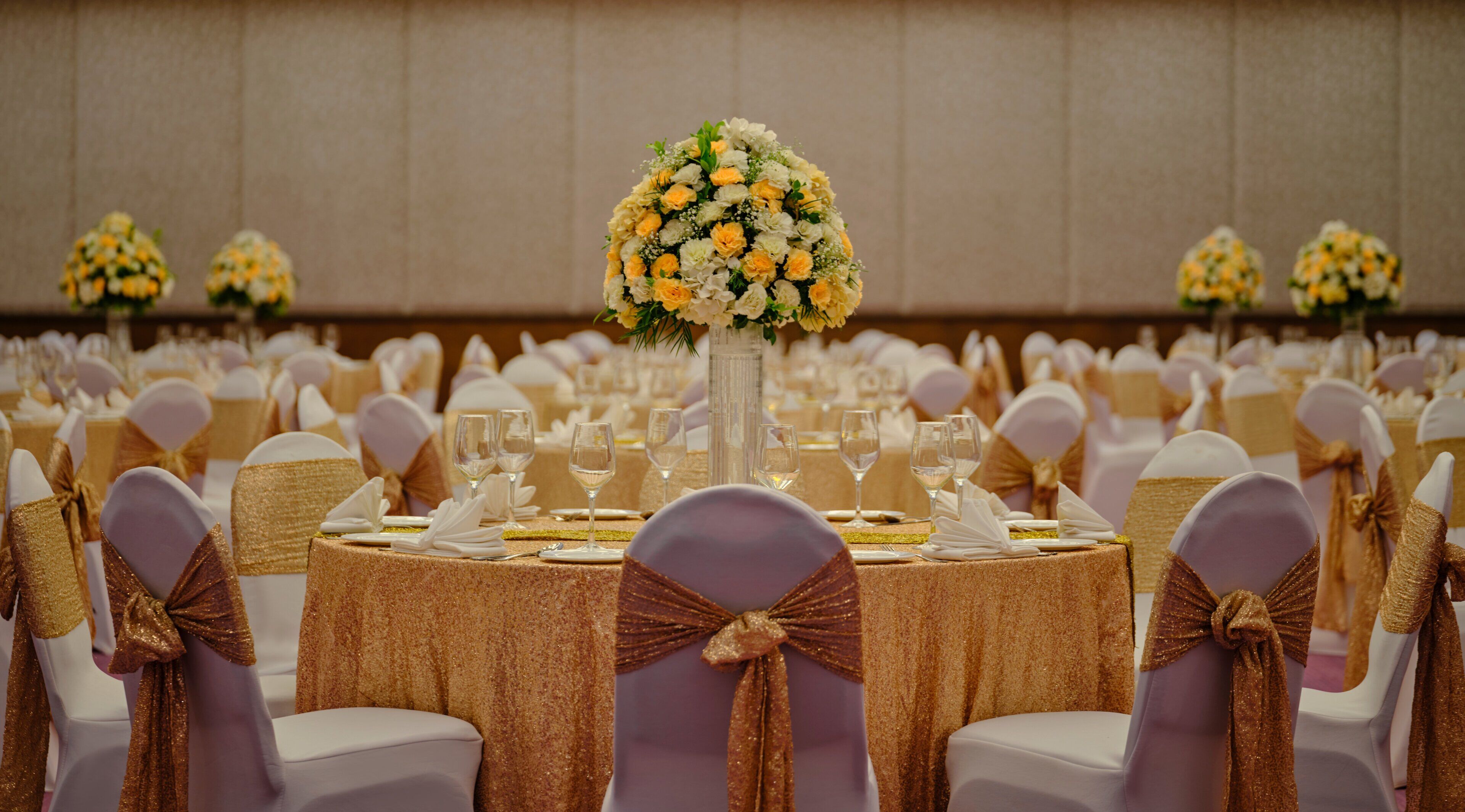 A banquet hall with round tables, white chairs with gold sashes, and floral centerpieces.