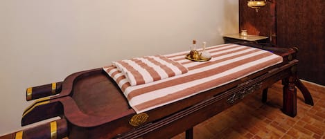 A wooden massage table with a striped towel, a small table with a lamp, and a wall-mounted scale.