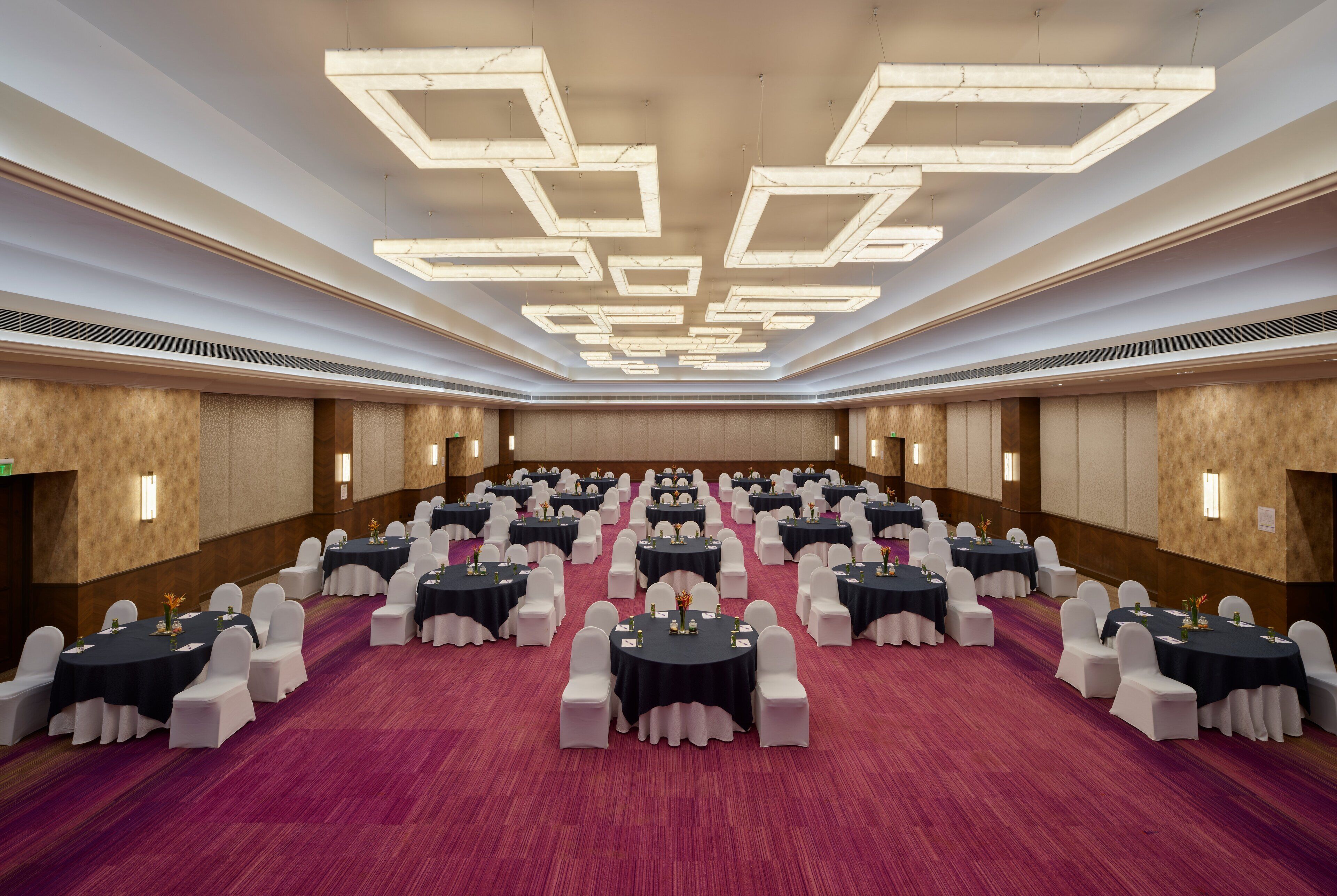 Un amplio salón de conferencias con varias mesas redondas, sillas blancas y una alfombra roja.