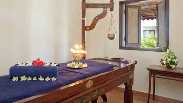Een houten massagetafel met een blauwe handdoek en decoratieve bloemen, een hanglamp en een klein tafeltje met een plant.