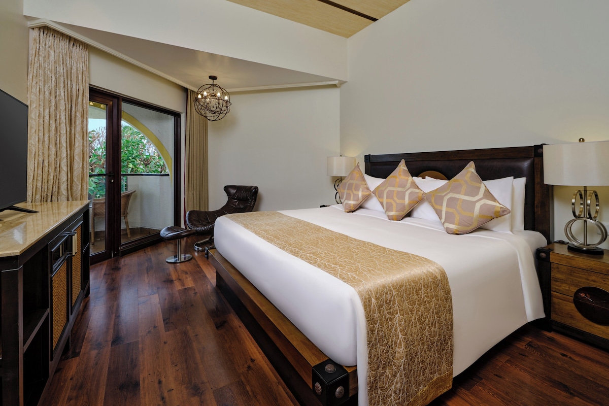 A hotel room with a large bed, a TV, a desk, and a view of greenery through a sliding door.