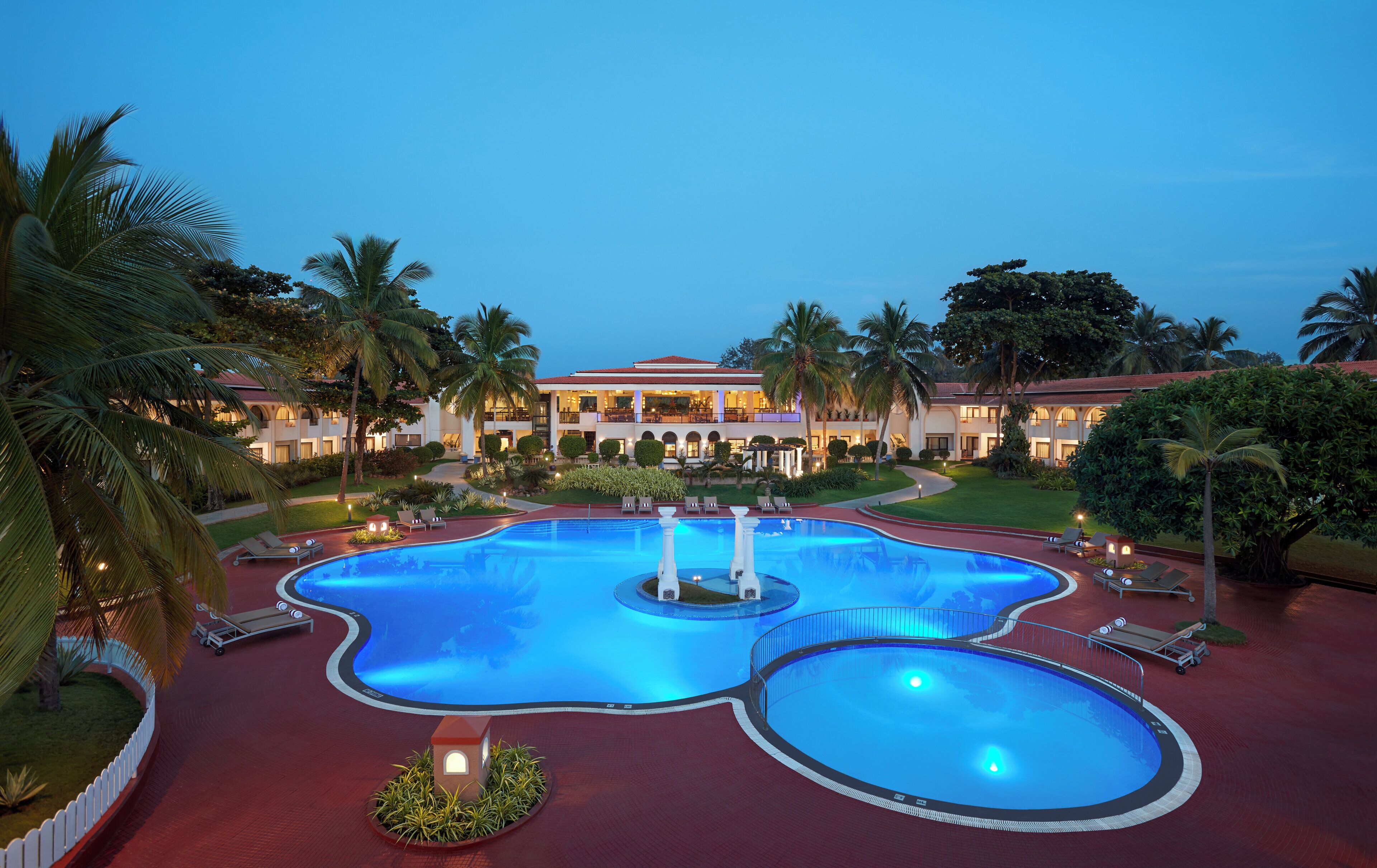 Um hotel com uma piscina grande, palmeiras e um edifício bem iluminado.