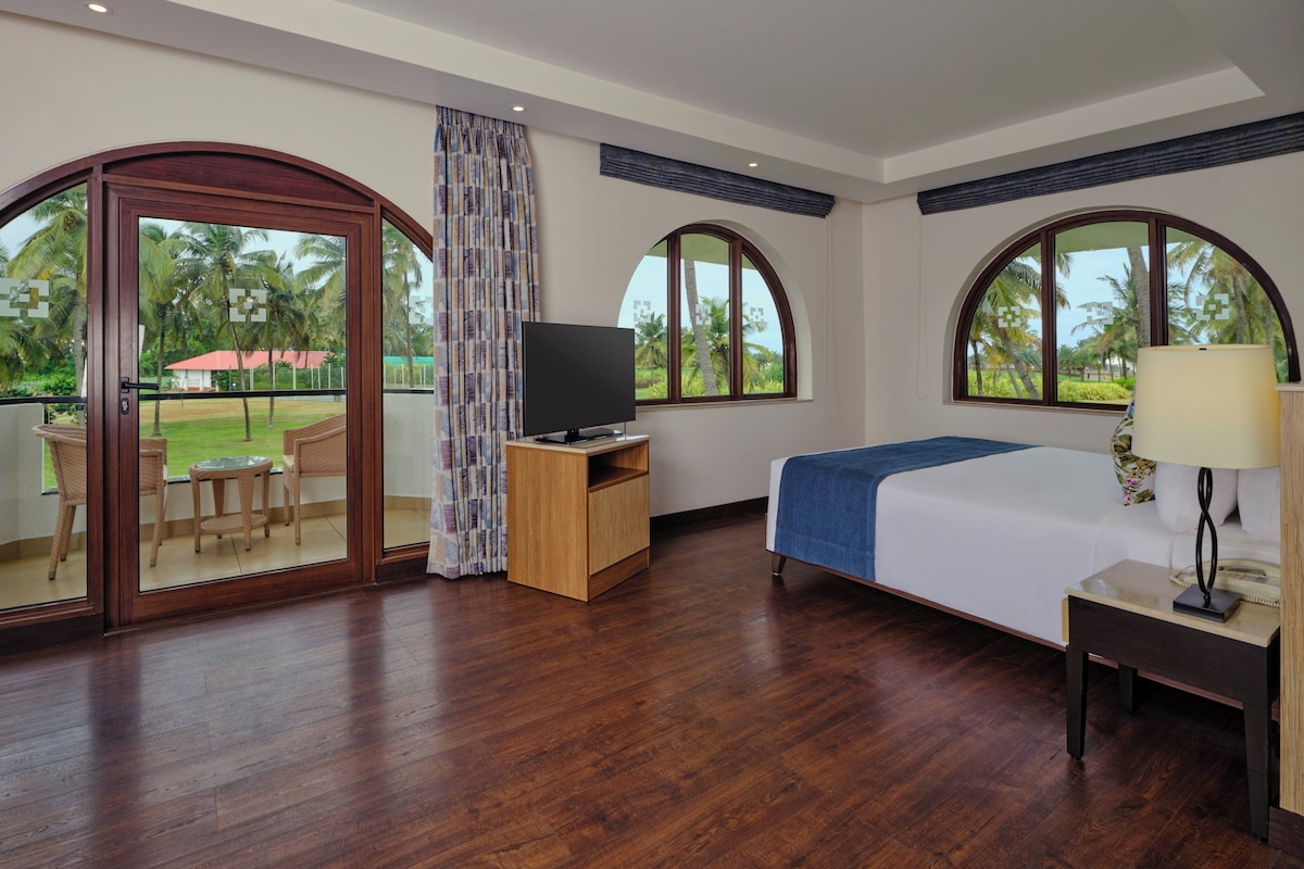 A hotel room with a wooden floor, a bed with a blue blanket, a TV on a wooden stand, a desk with a lamp, and large arched windows with a view of palm trees.