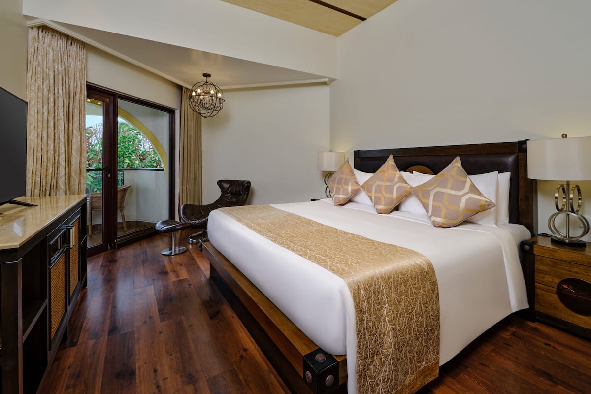 A hotel room with a large bed, a TV, a desk, and a view of greenery through a sliding door.