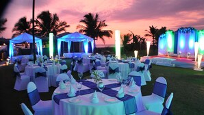 An outdoor event space with round tables set for dining, decorated with white tablecloths and blue sashes, and illuminated by ambient lighting.