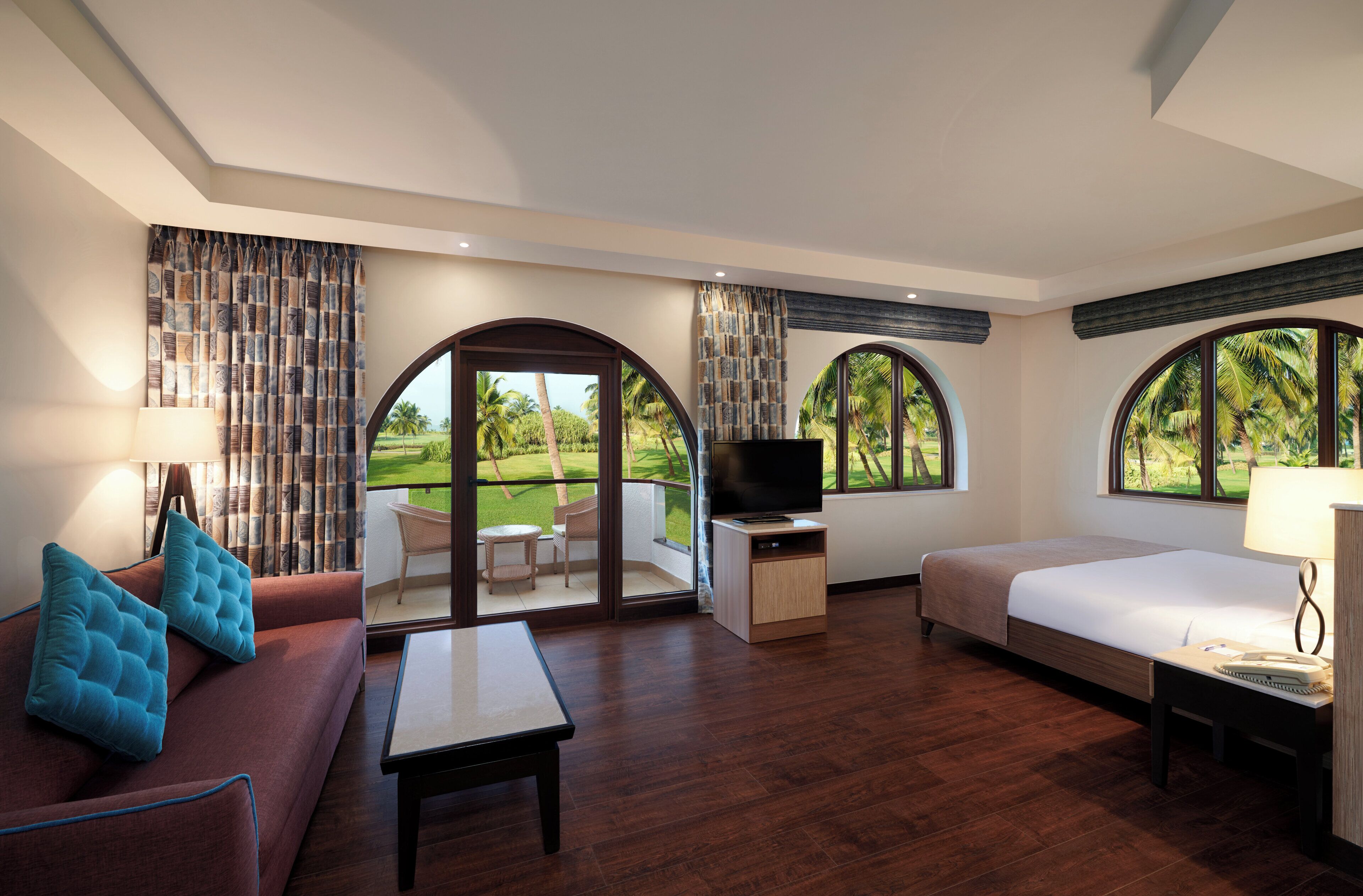 Habitación de hotel con sofá, cama, televisor, una mesita y balcón con vista a palmeras.