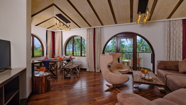 A spacious living area with wooden flooring, a dining table set for four, a large curved armchair, and a sofa. - Holiday Inn Resort Goa by IHG (Cavelossim)