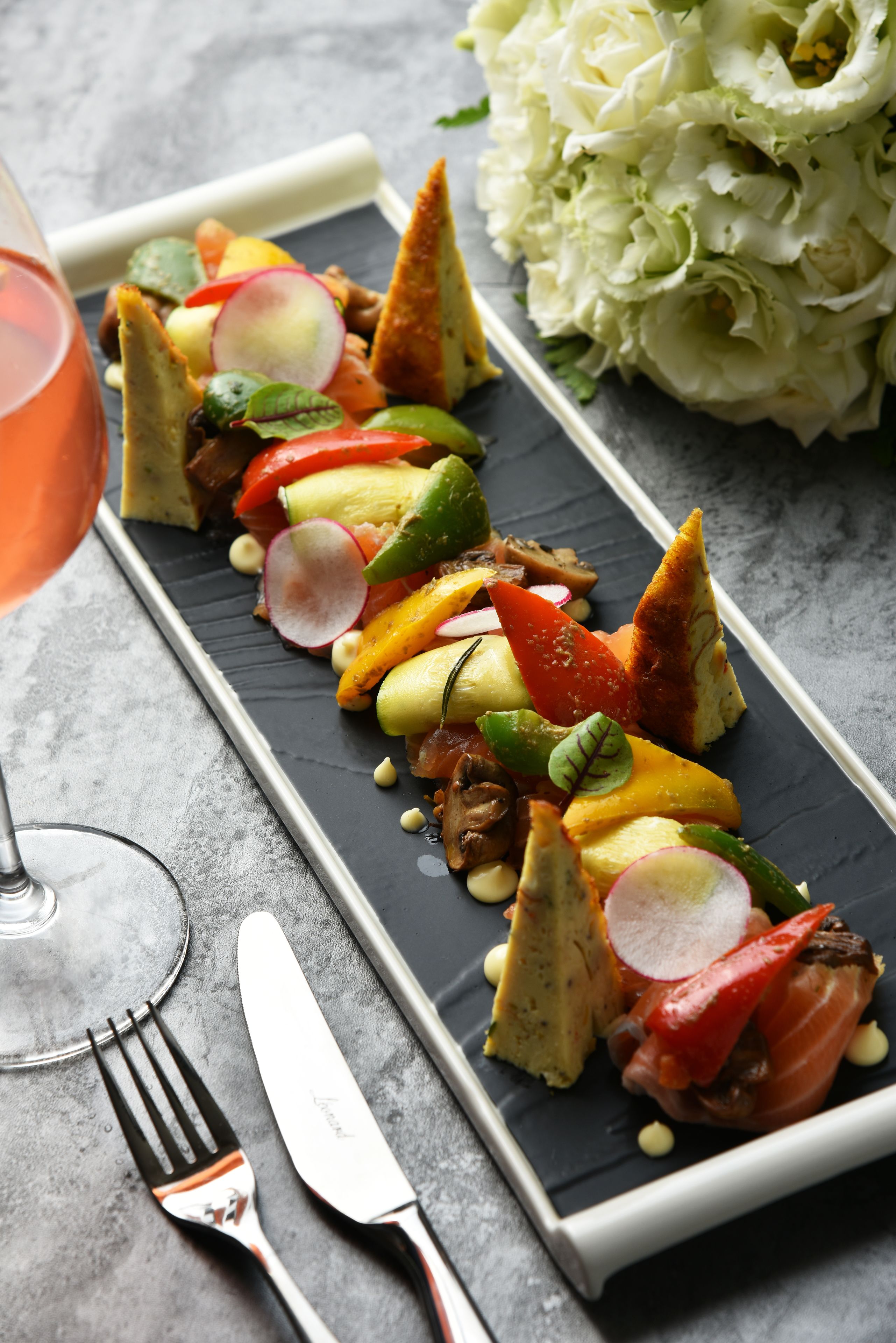 A rectangular plate with a variety of food items including vegetables, cheese, and meat, accompanied by a glass of drink and cutlery on a grey surface.