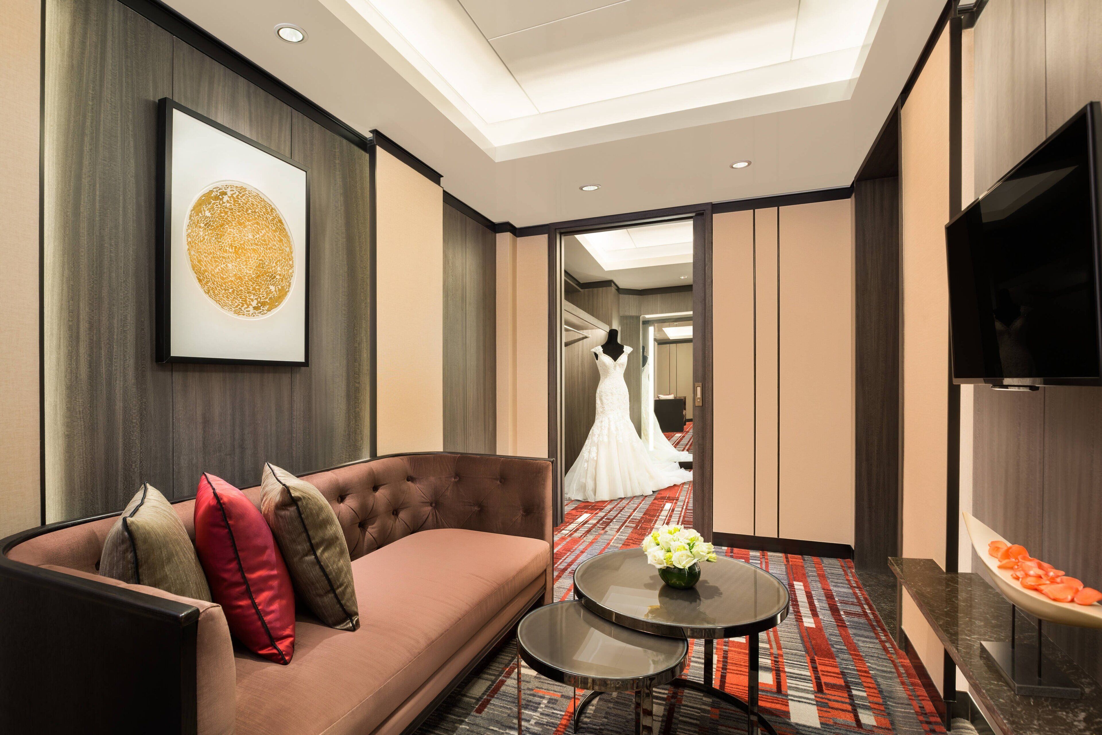 A modern hotel room with a sofa, a small round table with a vase, a flat-screen TV, and a large framed artwork on the wall.