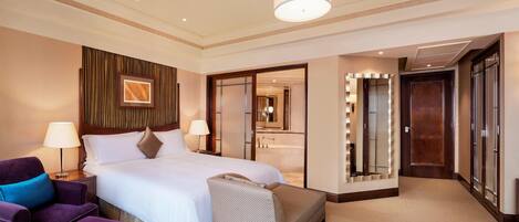 A hotel room with a large bed, two bedside tables with lamps, a purple armchair, a small sofa, a vanity with a mirror, and a door leading to a bathroom.