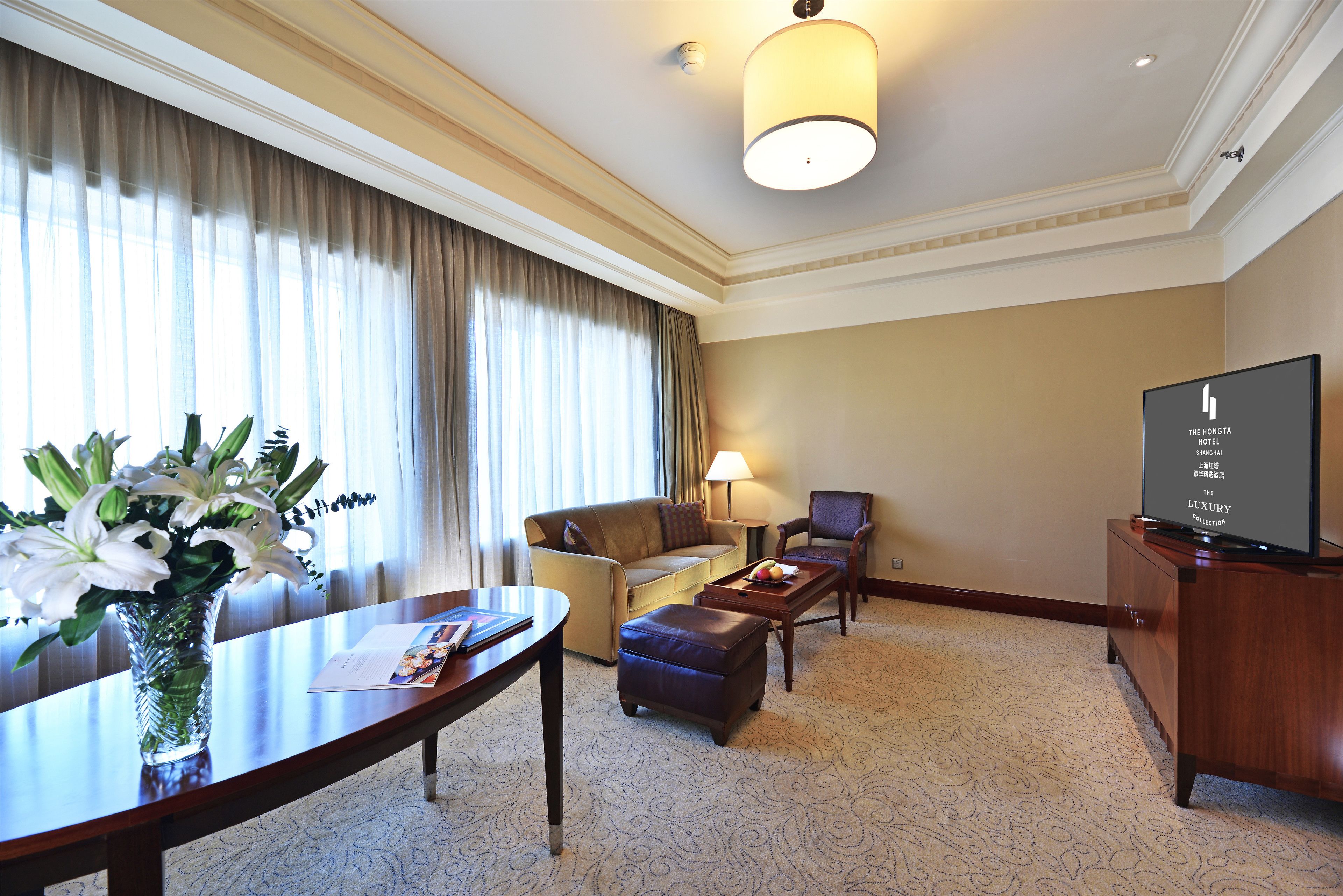 A hotel room with a sofa, armchair, ottoman, a dining table with a vase of flowers, a flat-screen TV, and a lamp.