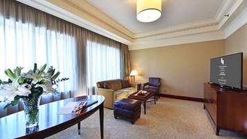 A hotel room with a sofa, armchair, ottoman, a dining table with a vase of flowers, a flat-screen TV, and a lamp.