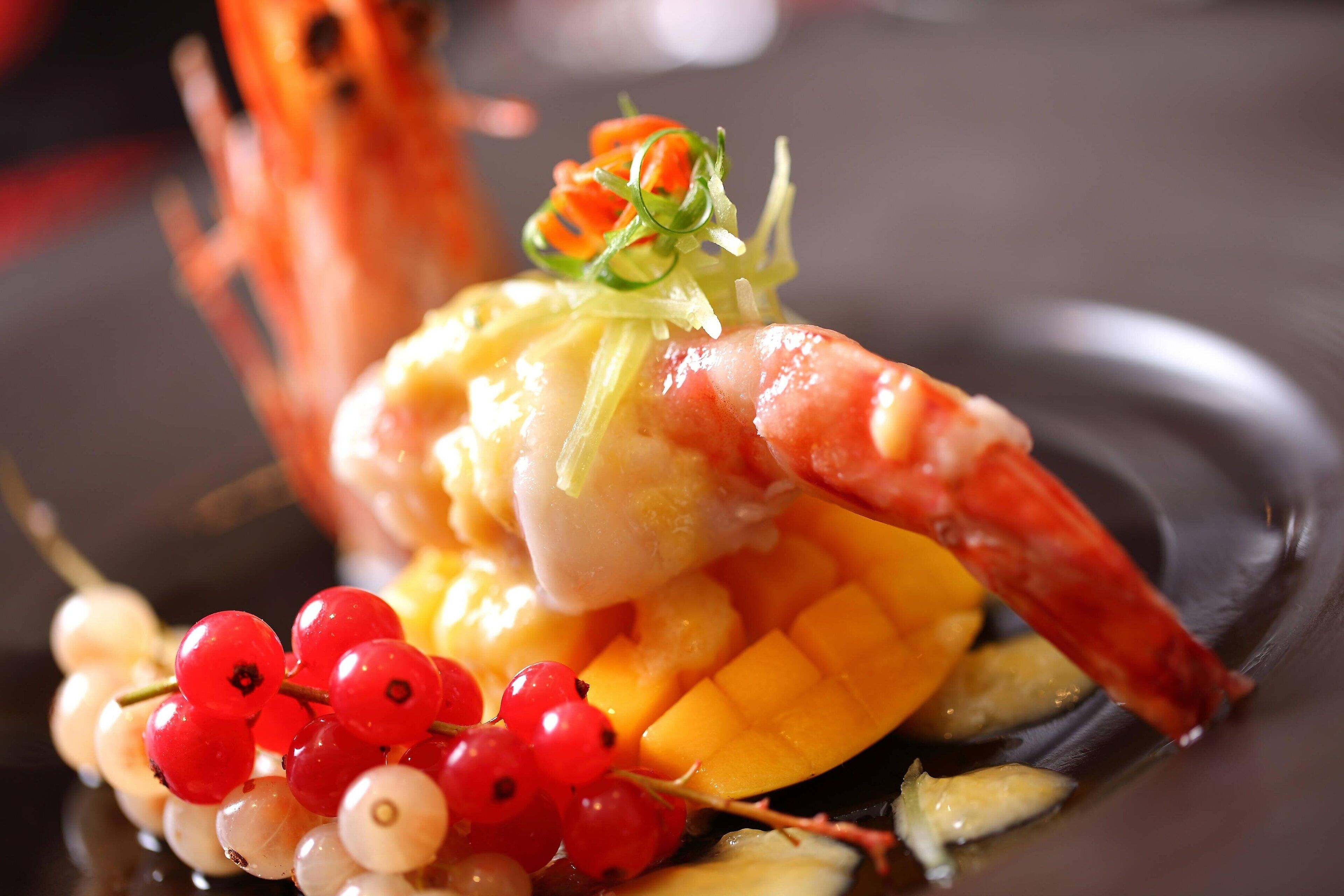 Un plat de fruits de mer composé de crevettes, de mangue et de groseilles rouges, servi dans une assiette noire.