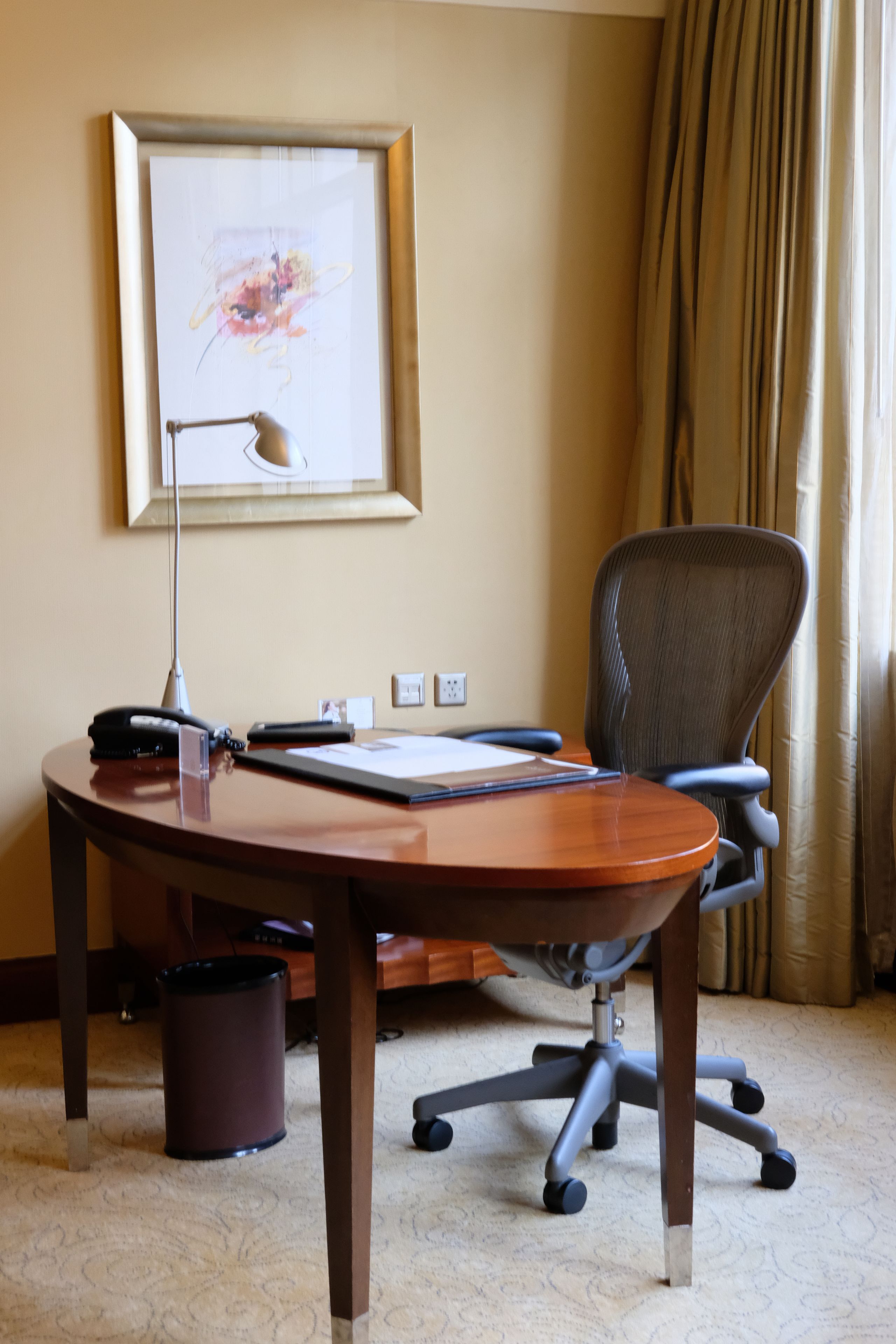 Un salón de conferencias con una mesa de madera, una silla de oficina negra, una lámpara de escritorio, una obra de arte enmarcada en la pared y un bote de basura.